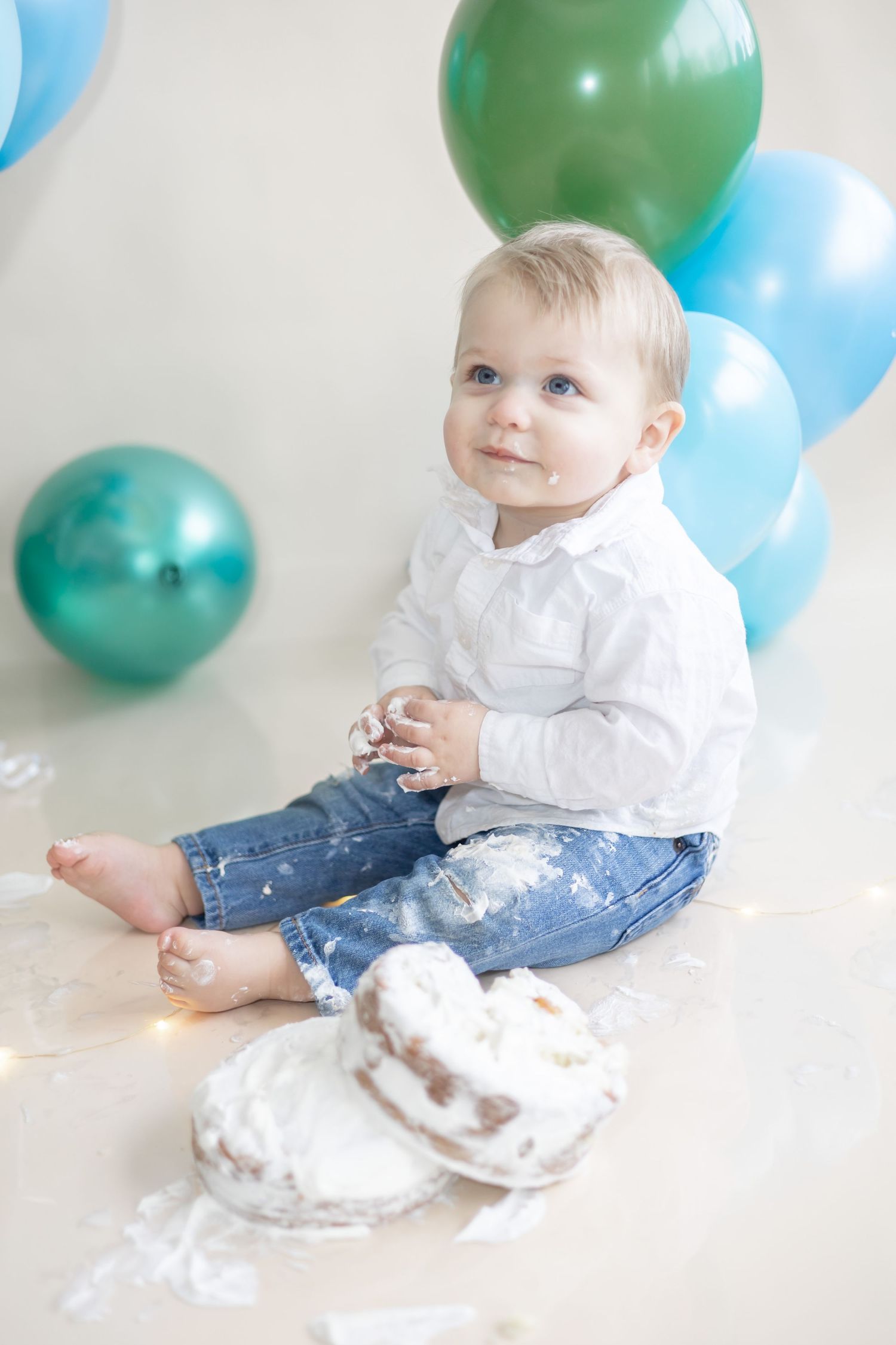 Baby in white shirt and jeans sits on floor with frosted cake and colorful party balloons in background.