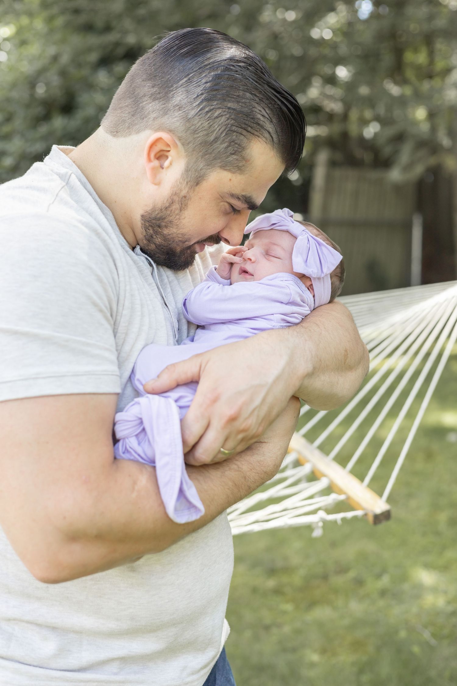 A tender moment shows a person in a white t-shirt holding and comforting a small baby in purple clothing outdoors by a hammock.