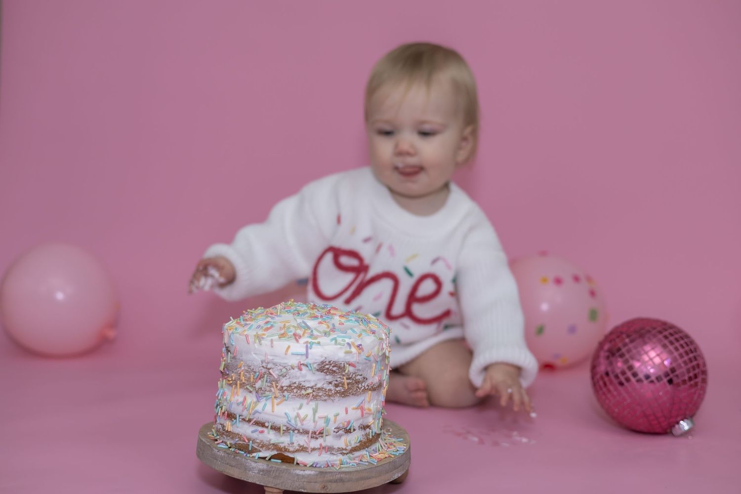 First birthday celebration with cake smash against pink backdrop with balloons and sparkly decorations.