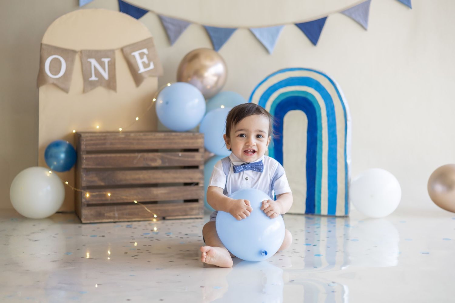 First birthday party setup with blue and white balloons, wooden crate, and blue rainbow balloon arch decoration.