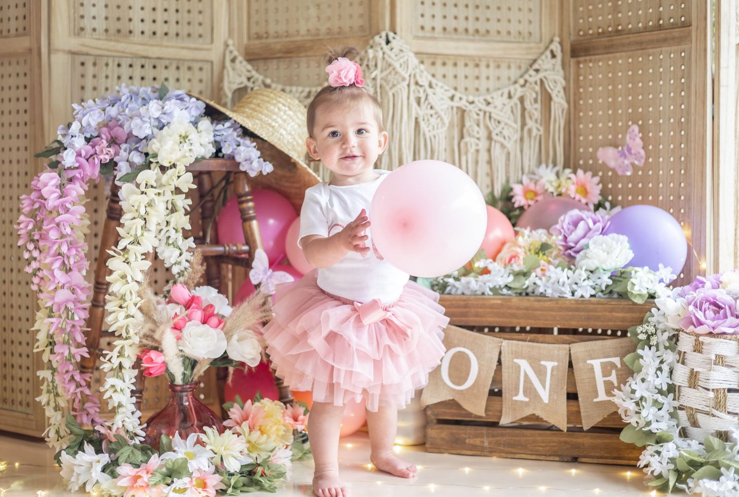 Baby in pink tutu celebrates first birthday with floral decorations, cotton candy and rustic 'ONE' banner backdrop.