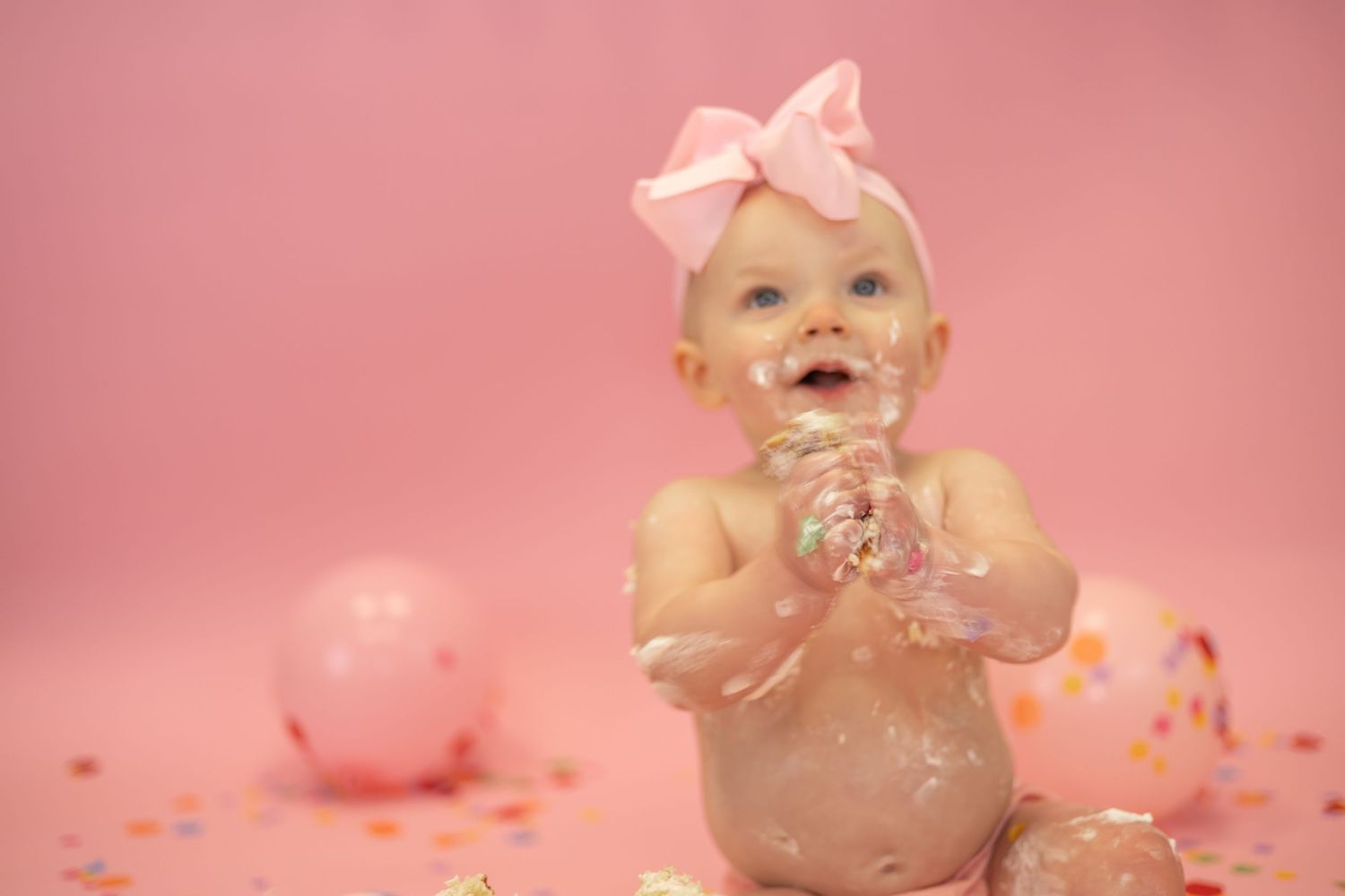 Baby wearing pink bow covered in glitter against pink backdrop with balloons and sparkles during cake smash photo session.