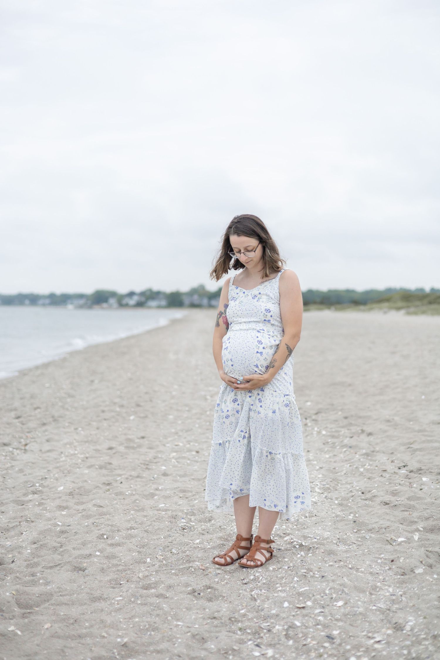 Mother-to-be cradles her baby bump while standing on a peaceful beach in a flowing white patterned sundress.