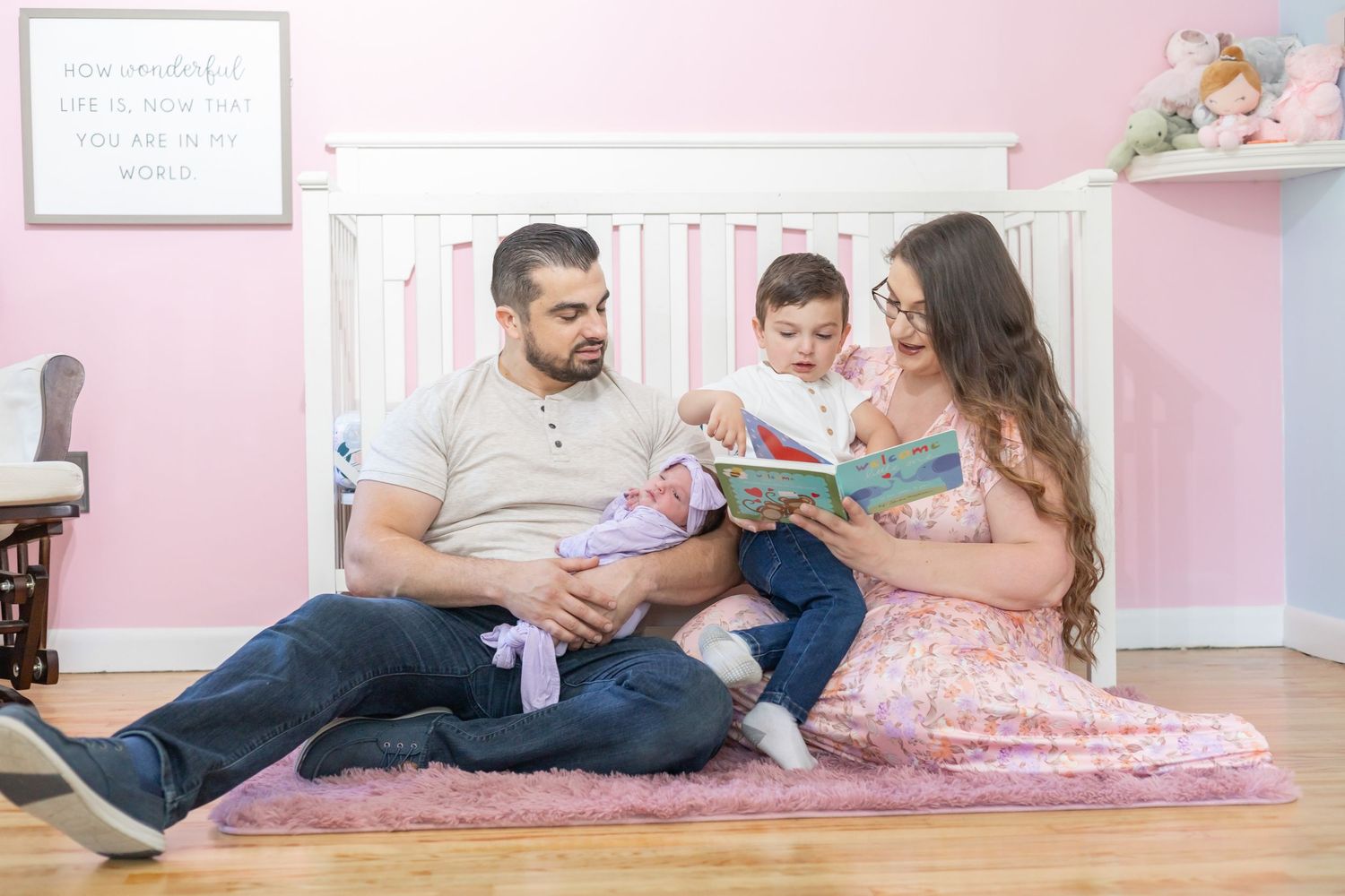Family reads together on a cozy pink floor pillow in a pink-walled nursery room.