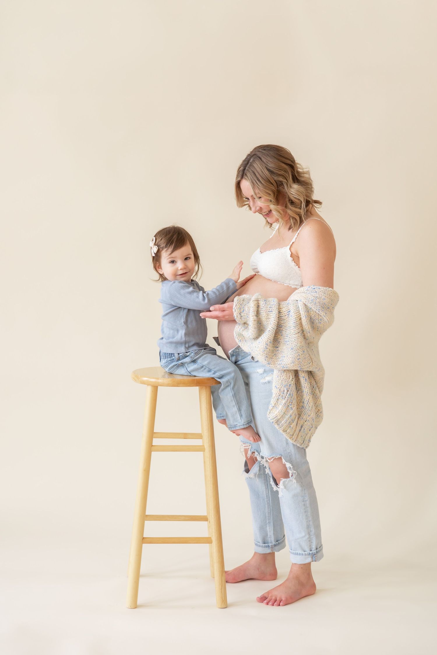 Mother and toddler share tender moment against beige backdrop as child sits on wooden stool in matching denim outfits.