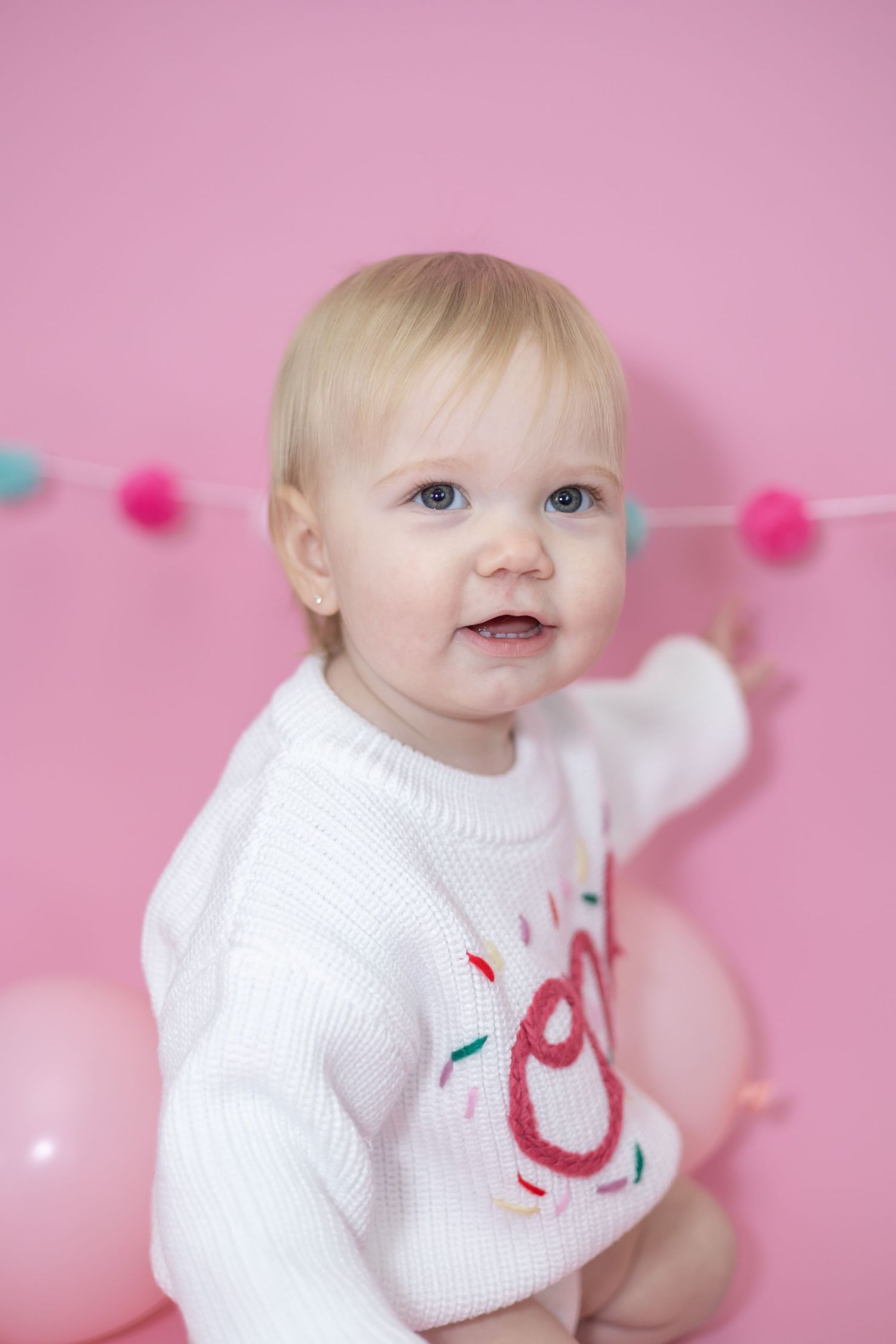 A small child wearing a white sweater smiles against a pink background with colorful pom pom decorations.