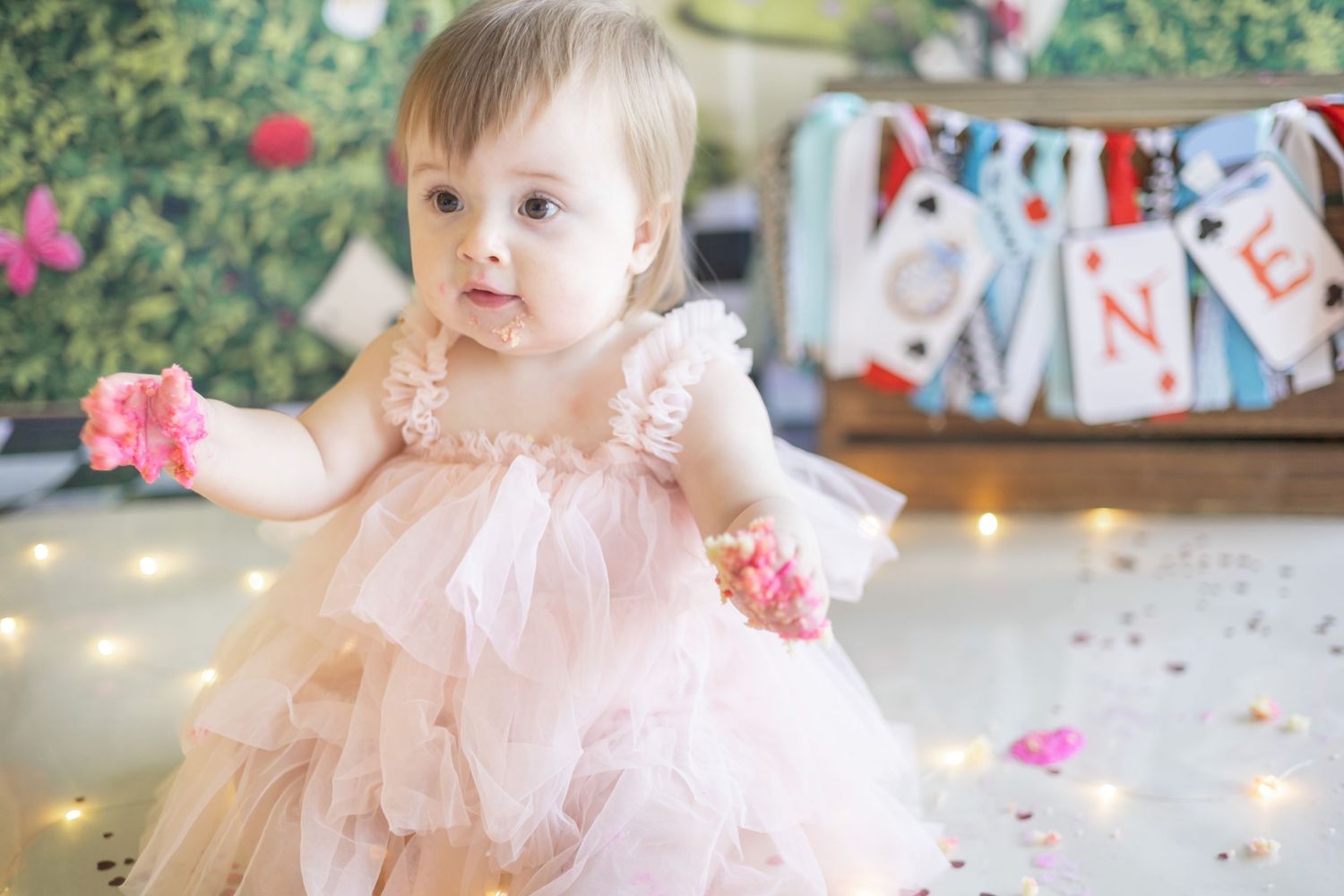 A small figure in a pink tulle dress reaching for flowers with decorative banners and string lights in the background.