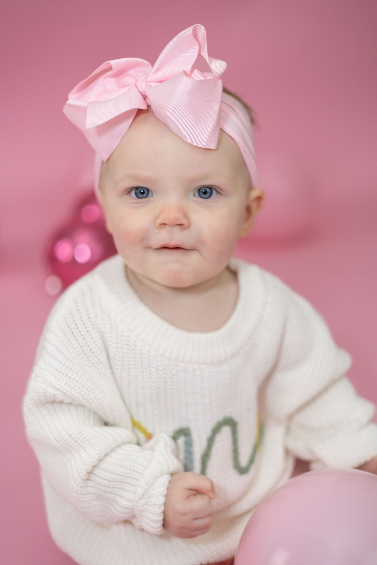 Baby wearing white sweater and pink bow headband against pink background with soft bokeh lights.