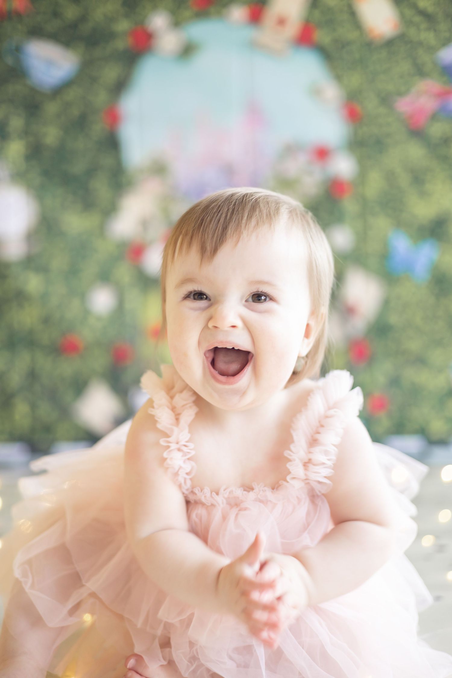 Happy baby wearing a light pink tulle dress smiles with pure joy against a soft bokeh Christmas tree background.