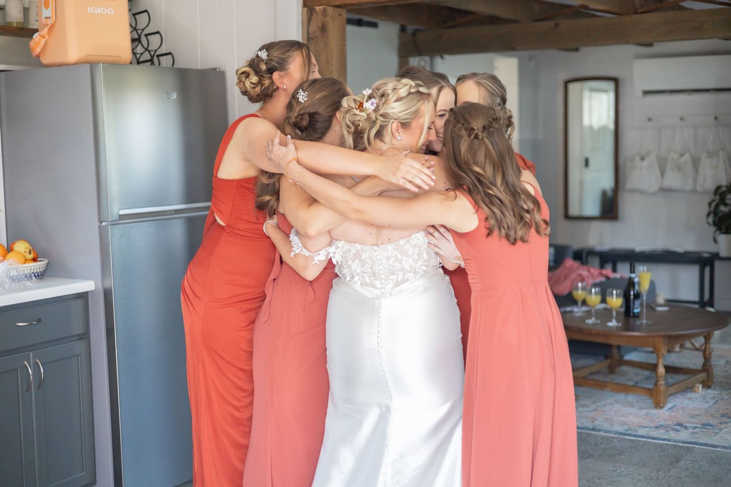 Wedding party members in coral dresses share emotional embraces during preparations before the ceremony.