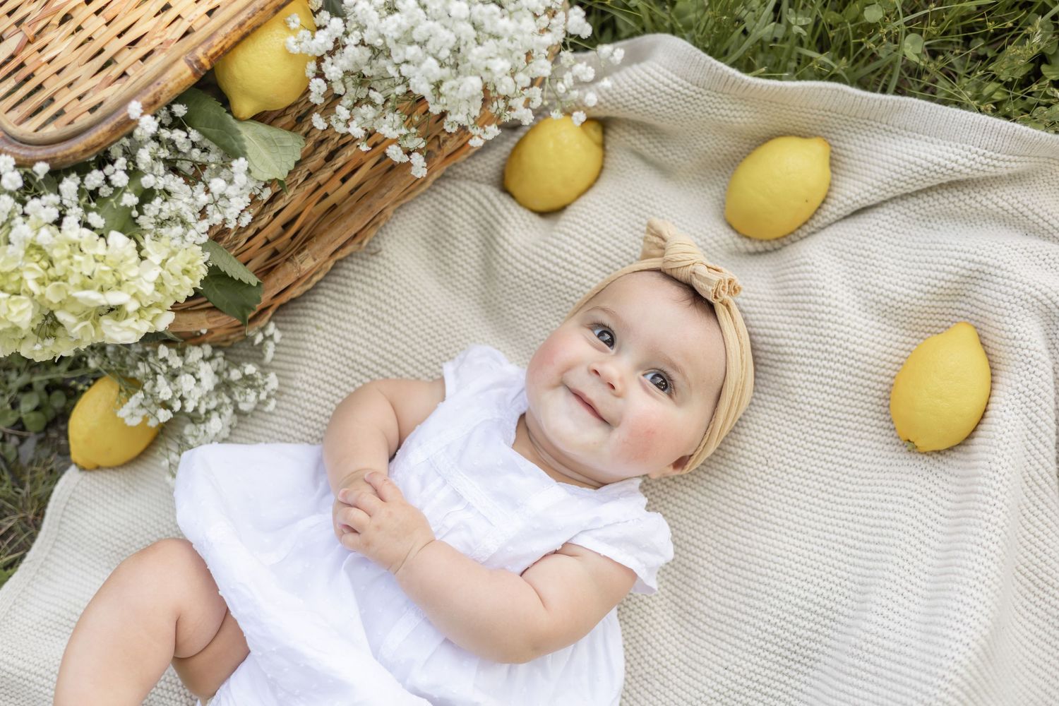 A white dress and bow on a blanket surrounded by fresh lemons and white spring flowers outdoors.