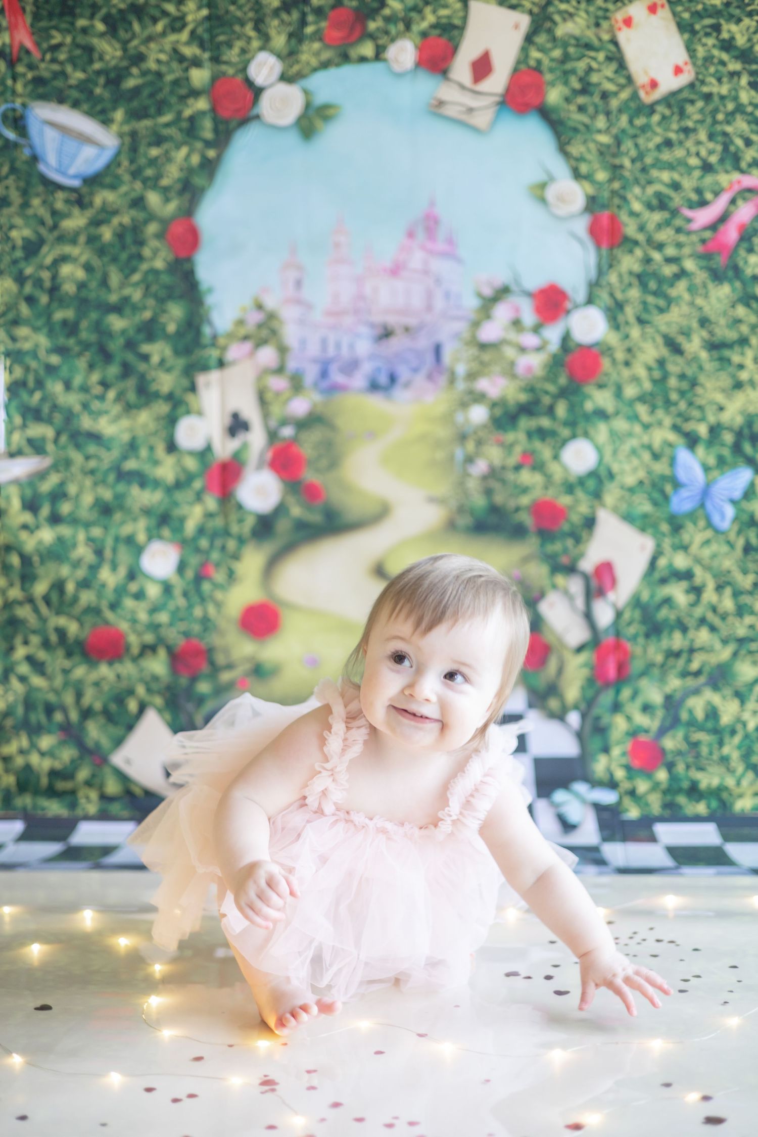 A magical wonderland scene with a pink castle backdrop, roses, playing cards, and whimsical decorations in Alice in Wonderland style.