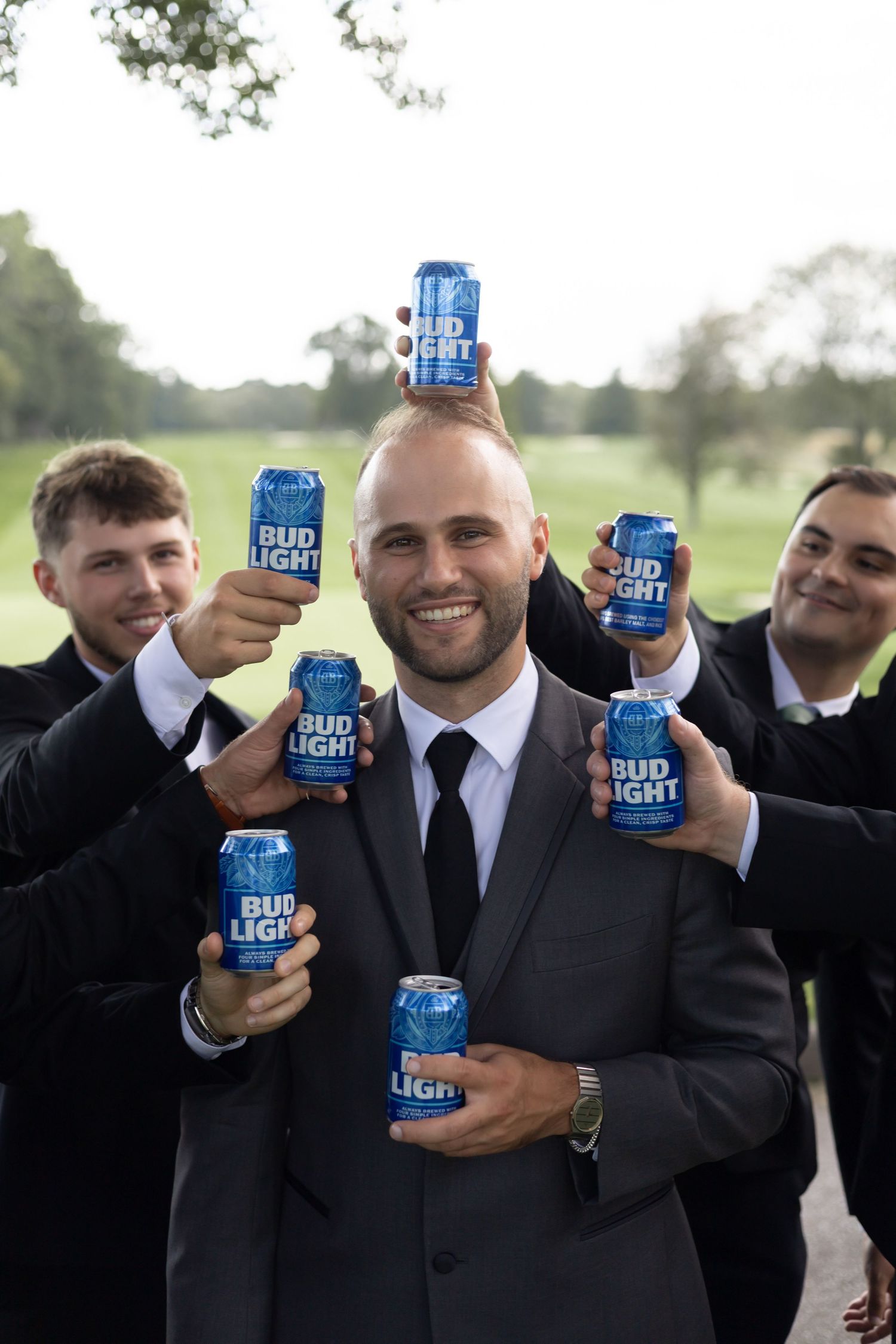Wedding party members in dark suits raise Bud Light beers in a celebratory toast outdoors.
