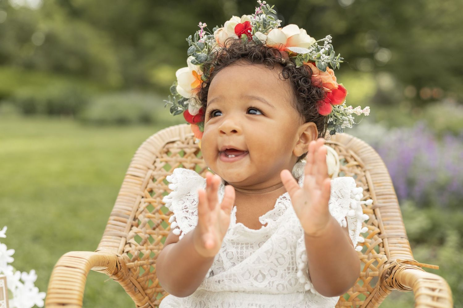 Joyful toddler wearing white lace dress and floral crown sits in wicker chair in garden setting, clapping hands.