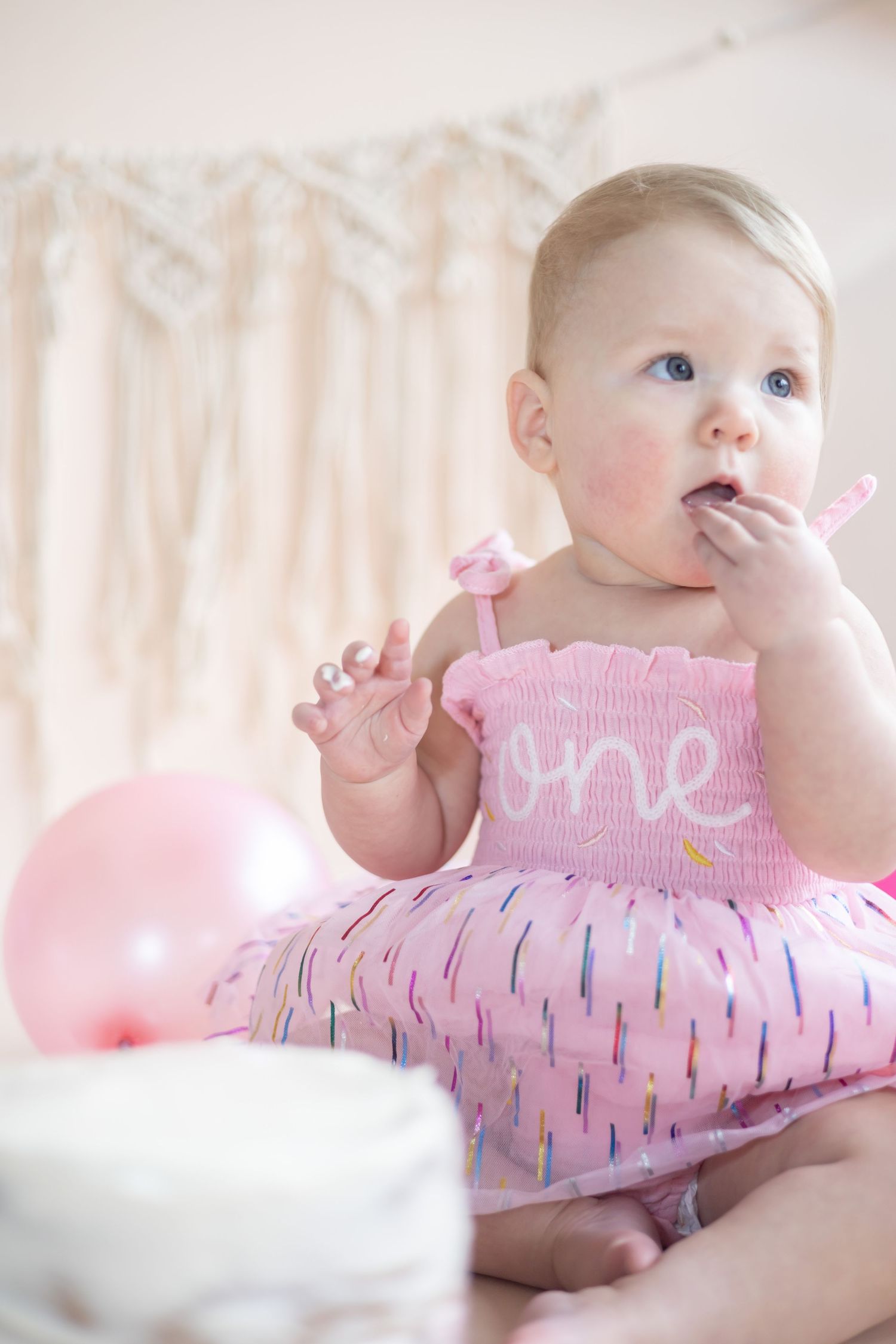 Baby in pink smocked dress with sprinkle pattern sits with balloon at first birthday celebration.