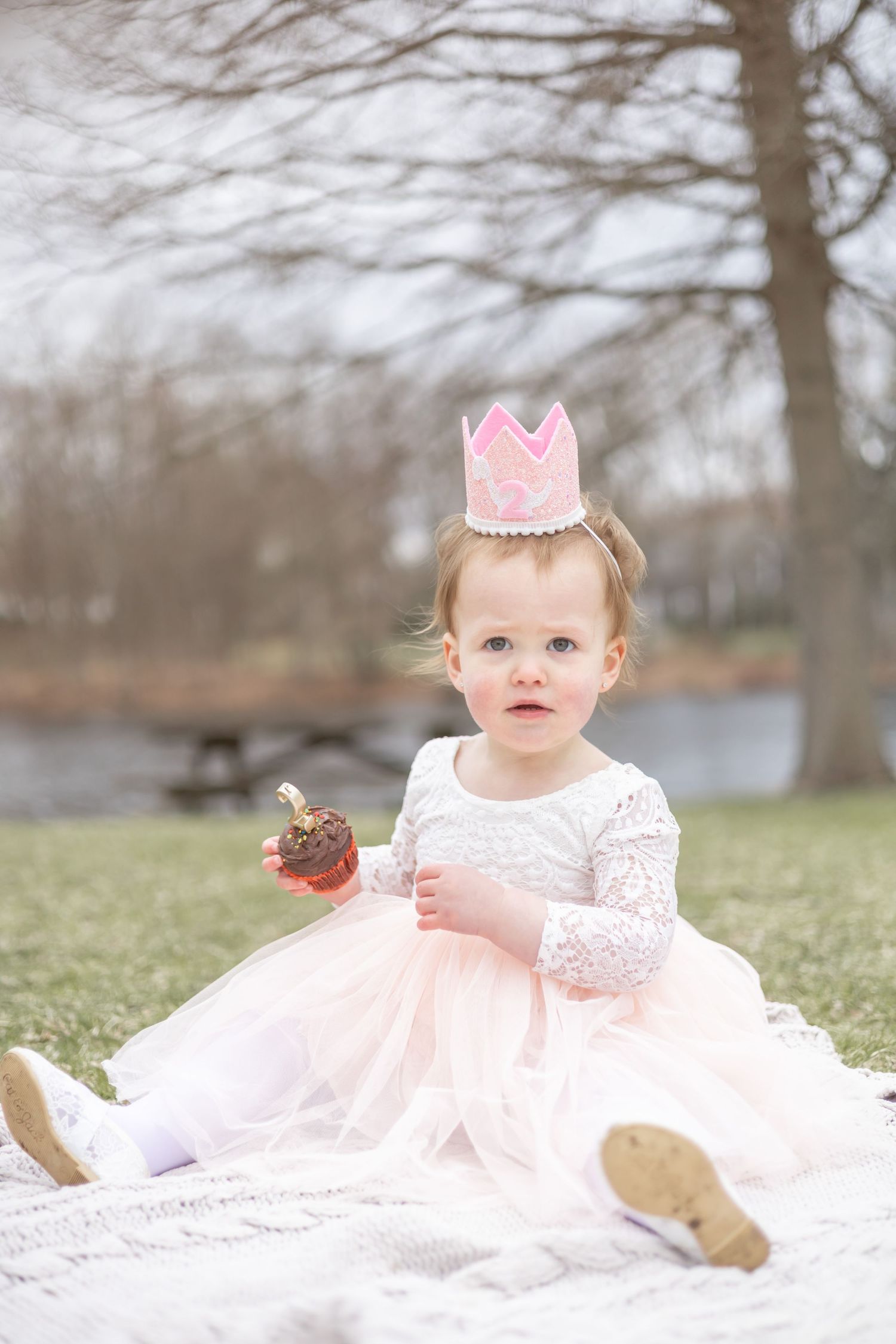 Baby in white lace dress and pink crown poses on blanket outdoors during springtime photo session.