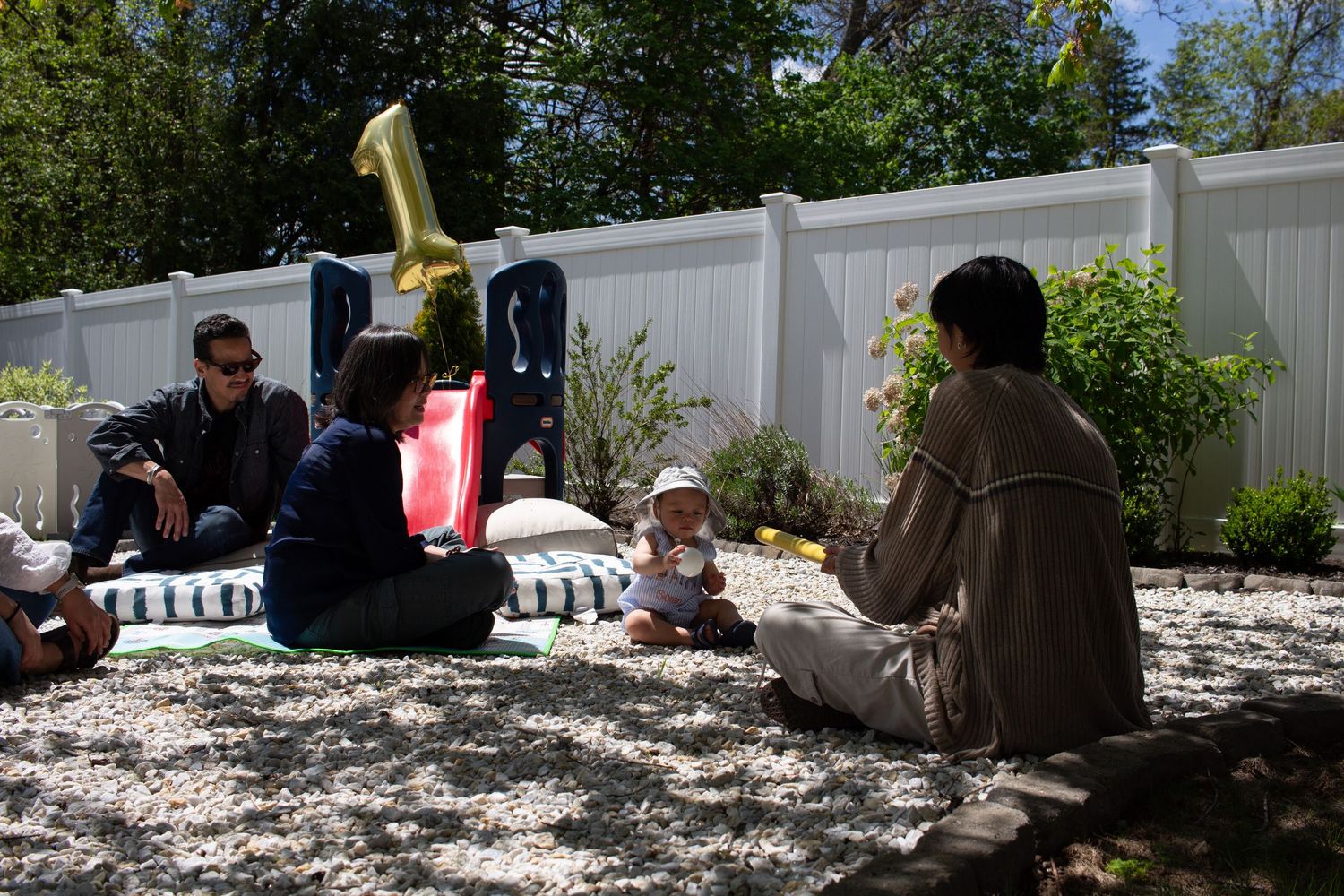 People gather outdoors for a casual first birthday celebration with party decorations near a white fence.