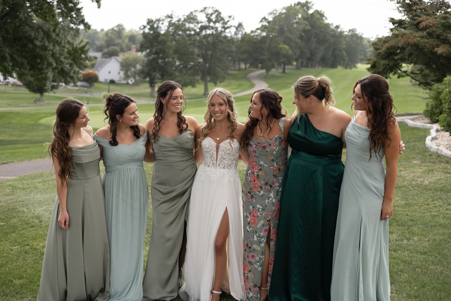 Group of bridesmaids in assorted blue, green and floral dresses standing together at an outdoor wedding venue.