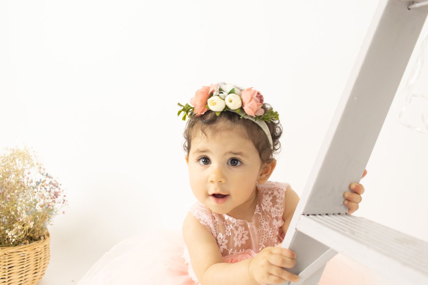 Sweet baby wearing floral crown sitting next to pink balloons in soft white studio setting for first birthday photoshoot.