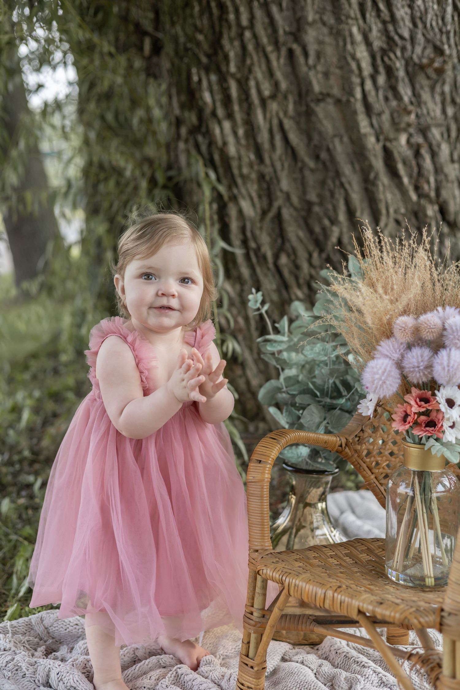 A toddler in a pink tulle dress stands by a wicker chair with dried floral arrangements under a large tree.
