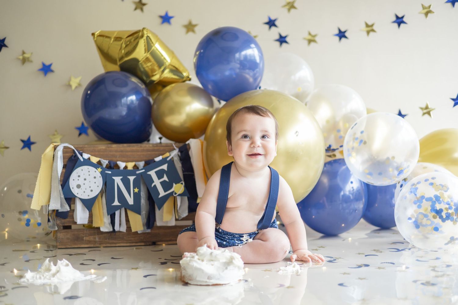 A first birthday cake smash setup with blue and gold balloons, a wooden crate, and festive party decorations.