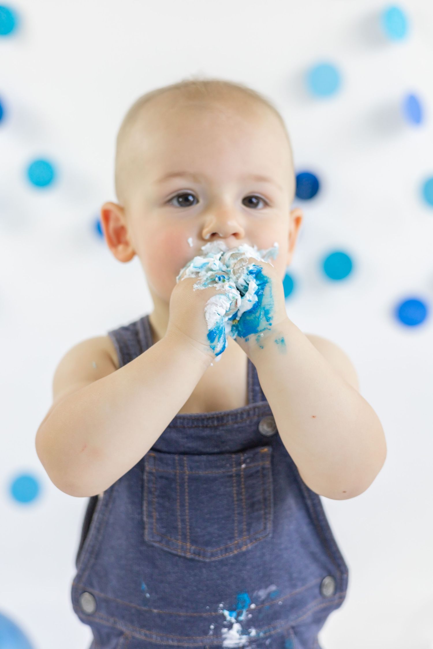 Baby in denim overalls gets messy eating blue frosting cake during cake smash photo session.