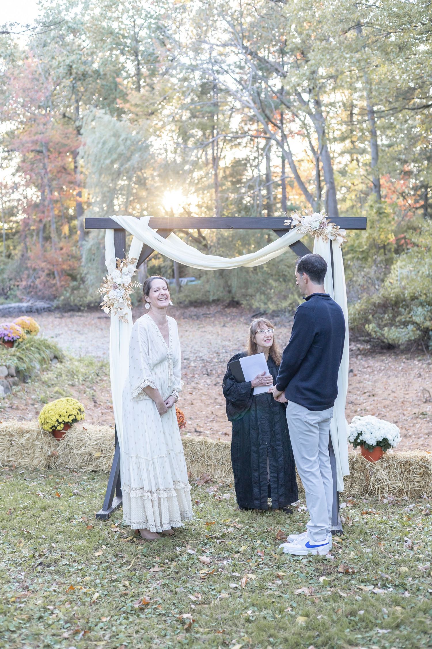 An outdoor wedding ceremony under a wooden arch decorated with white fabric and flowers during golden hour sunlight.