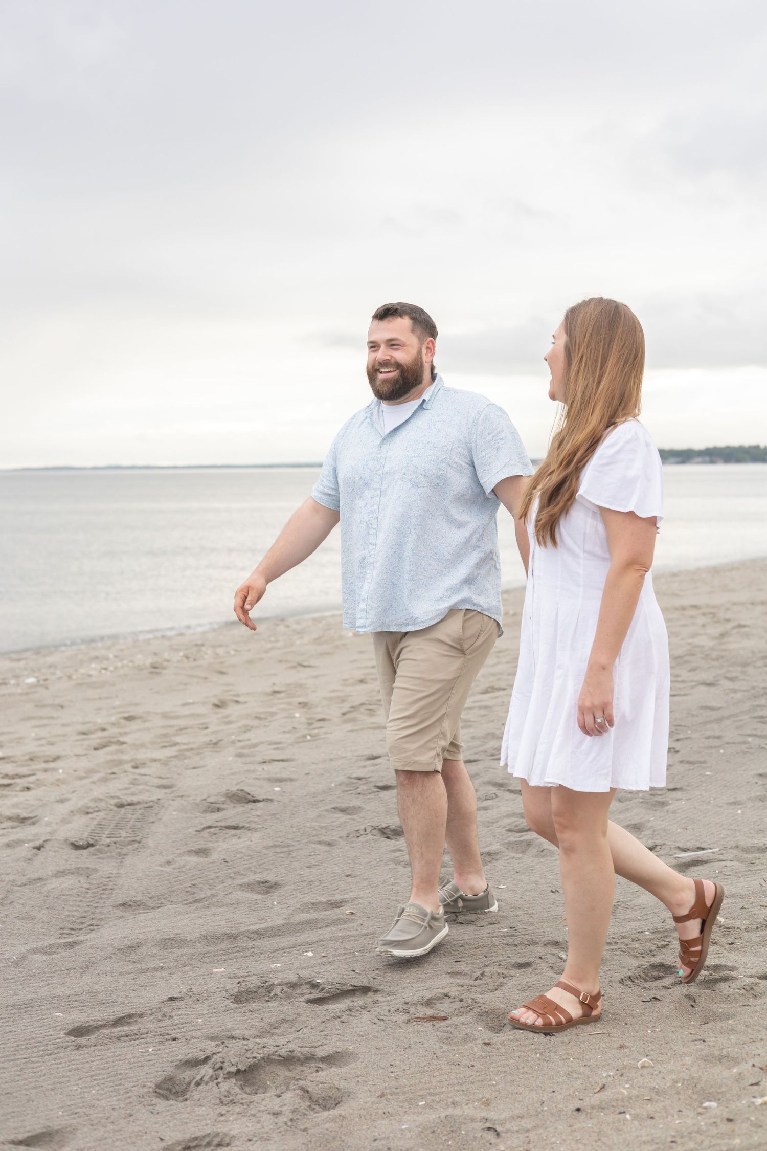 A couple in casual summer attire walking together on a sandy beach during an overcast day.