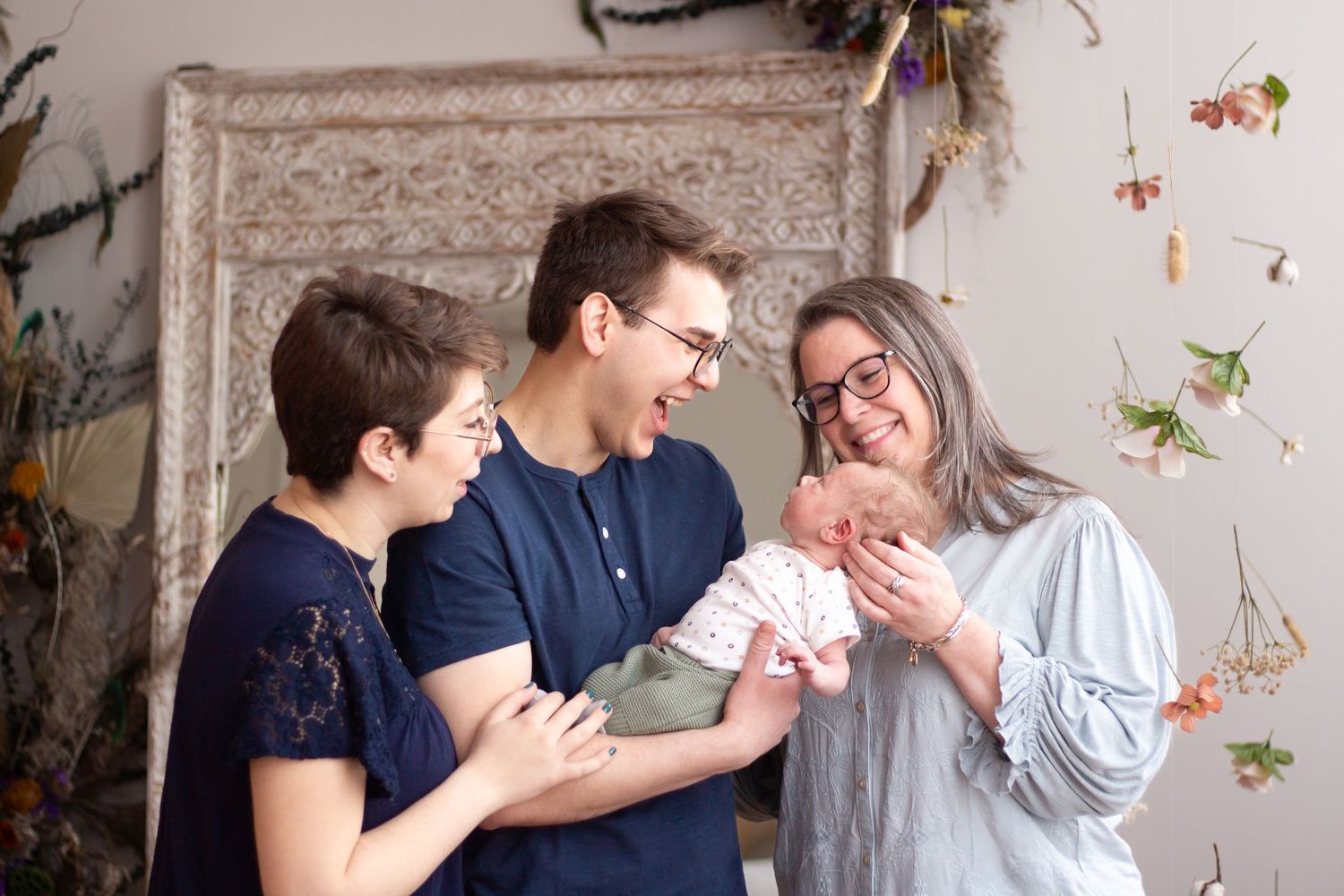 A group shares a happy moment together while holding a newborn baby in front of a floral wall backdrop.