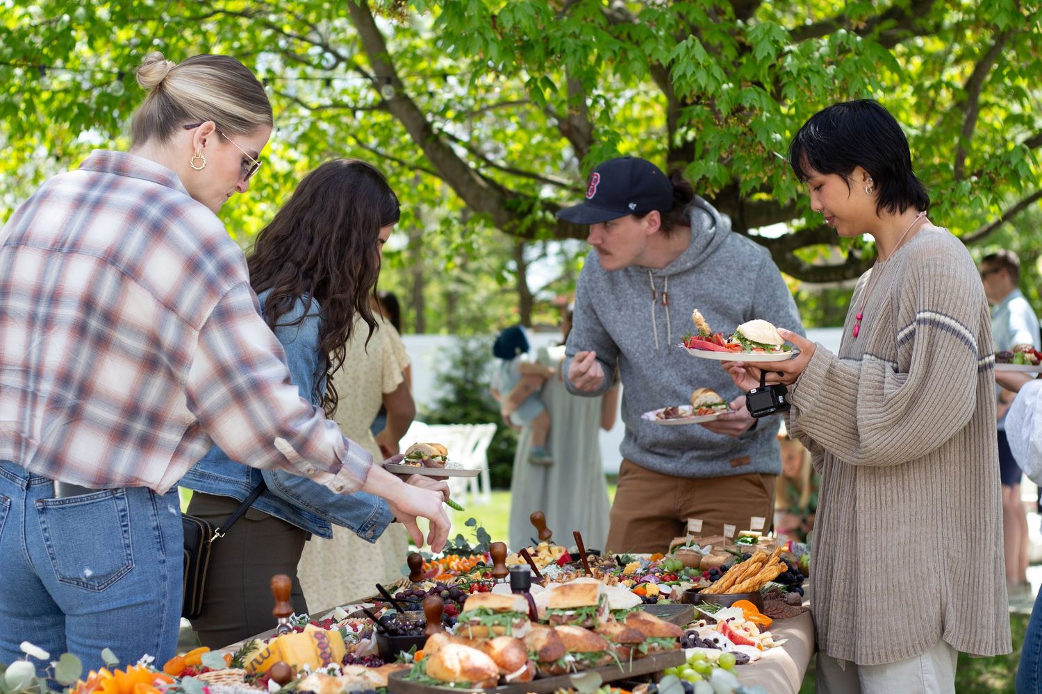 People enjoying food at an outdoor buffet table under green trees during a casual gathering.