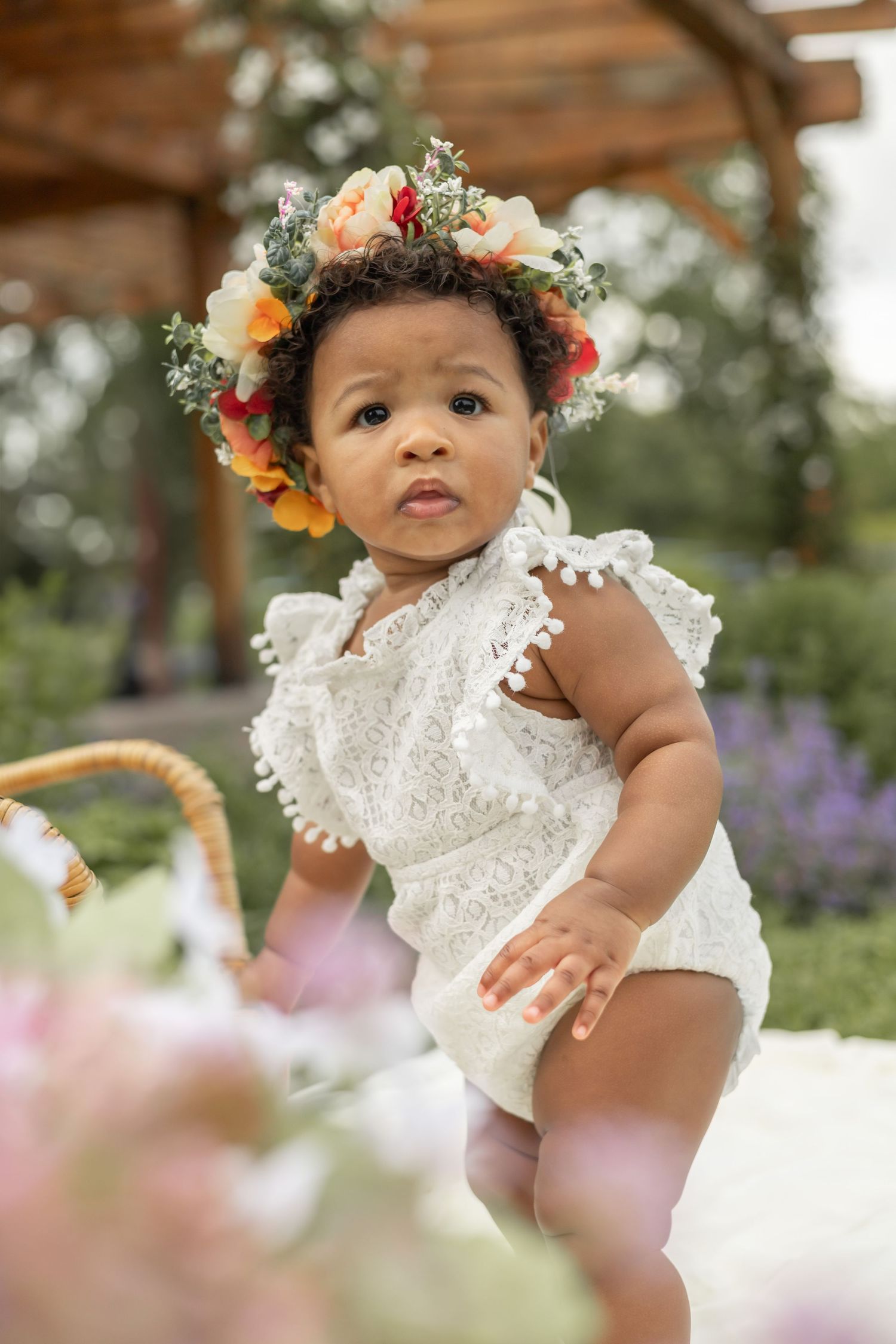 Baby in white lace romper and floral crown stands in garden with purple flowers in background.