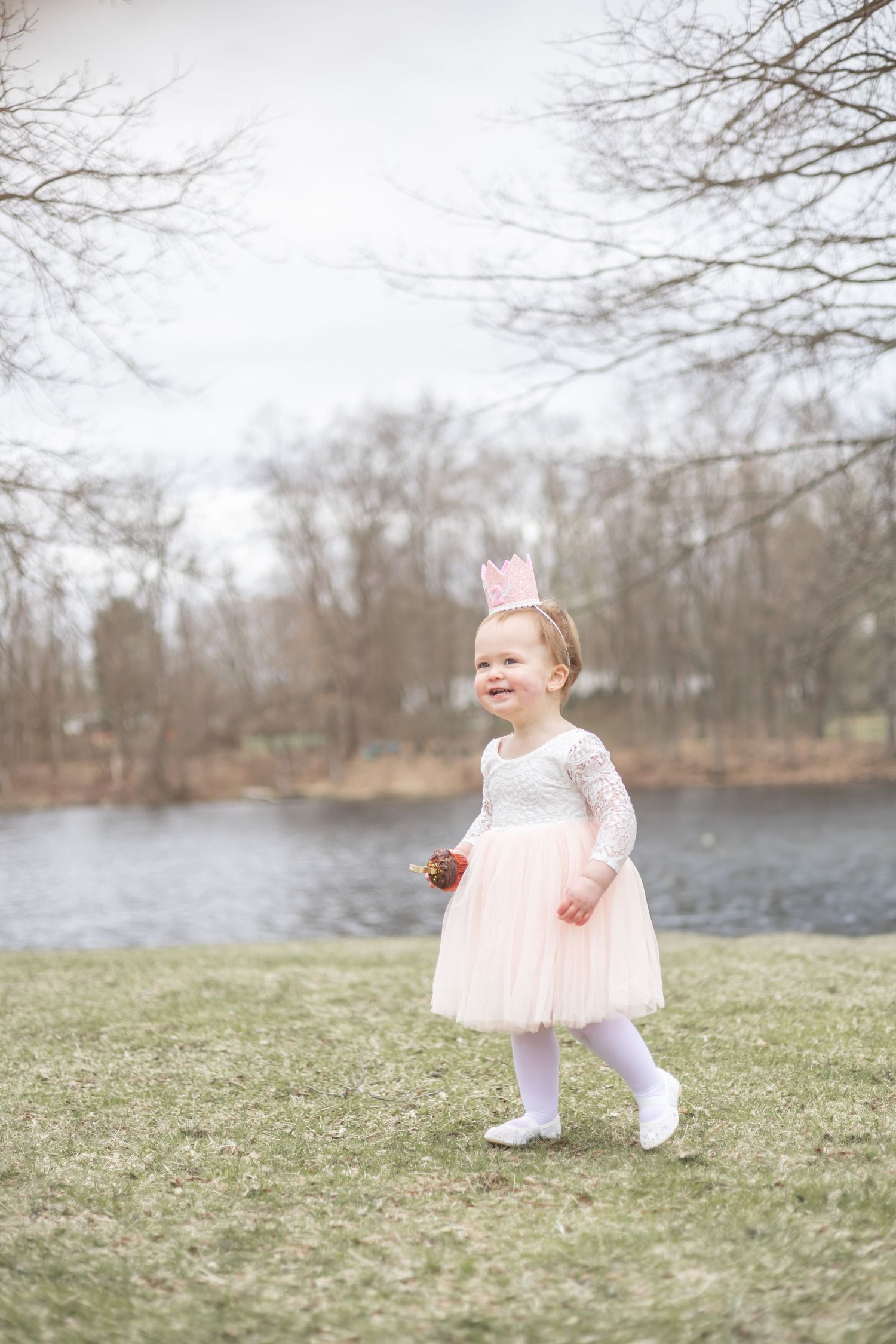 Small child wearing a pink tulle dress and bunny ears stands on grass by a lake on a misty spring day.