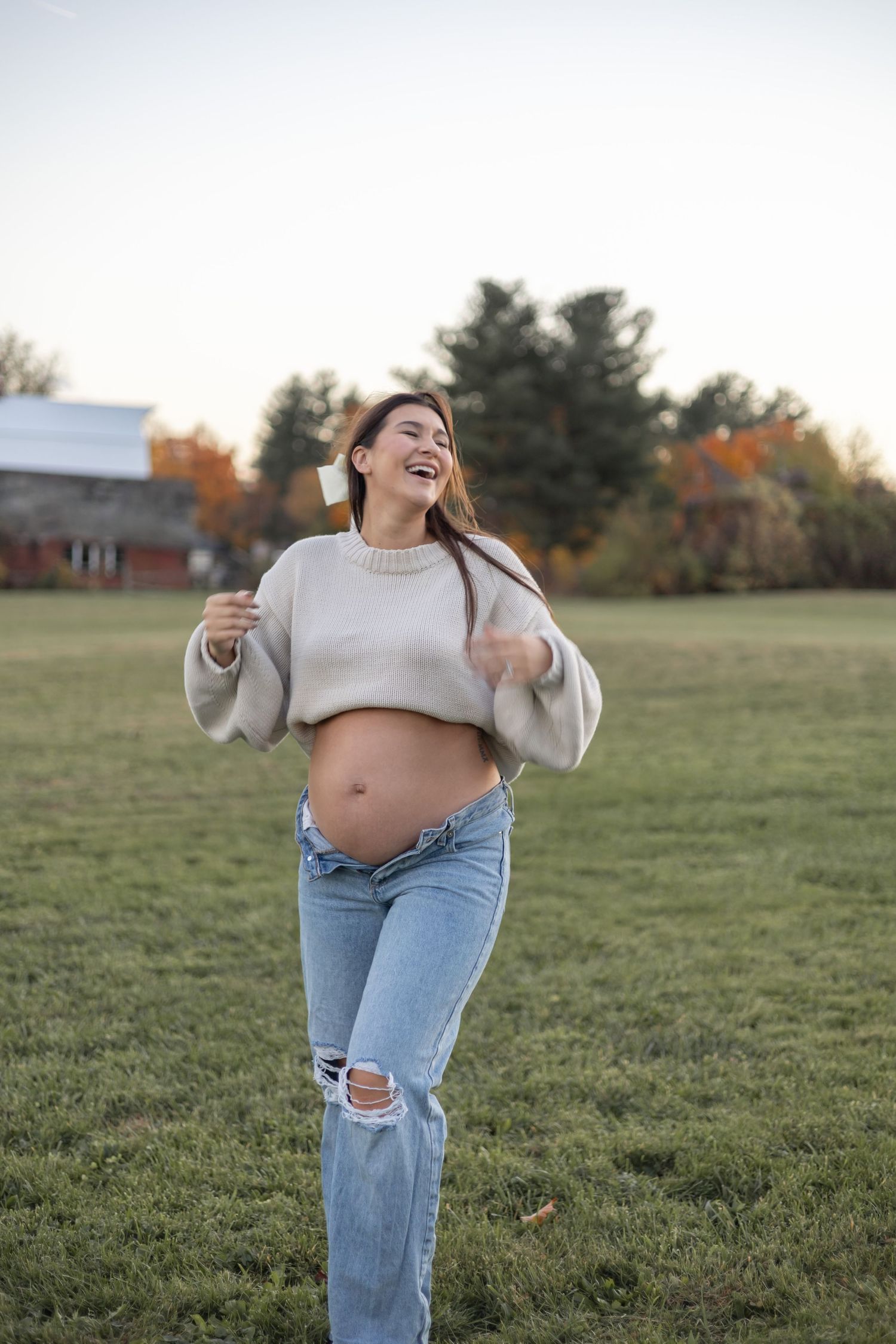 A joyful expecting mother in casual attire laughs while showing her baby bump in an autumn field at sunset.