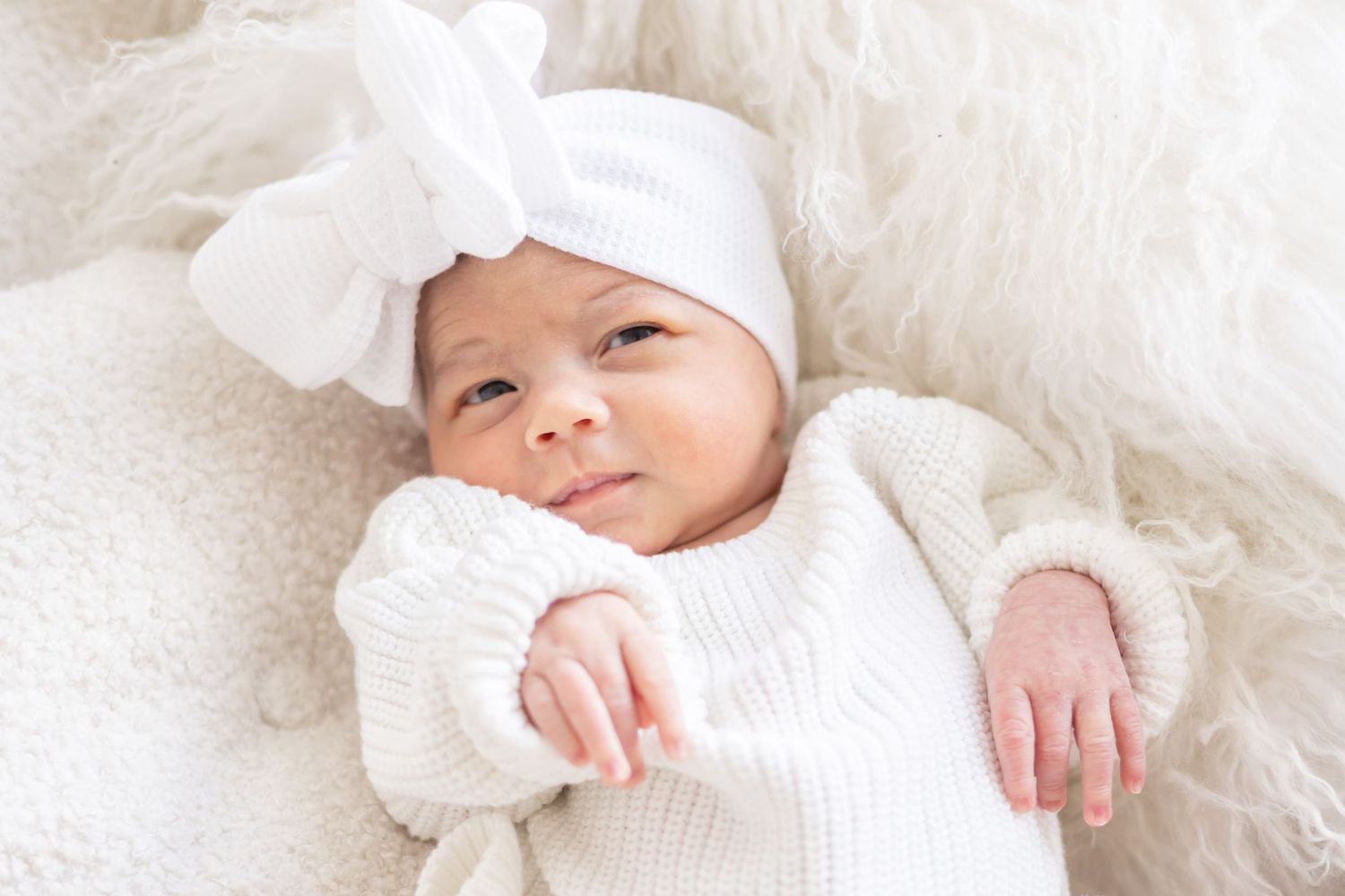 A baby in a white outfit lays on soft white bedding with their arms spread out comfortably.