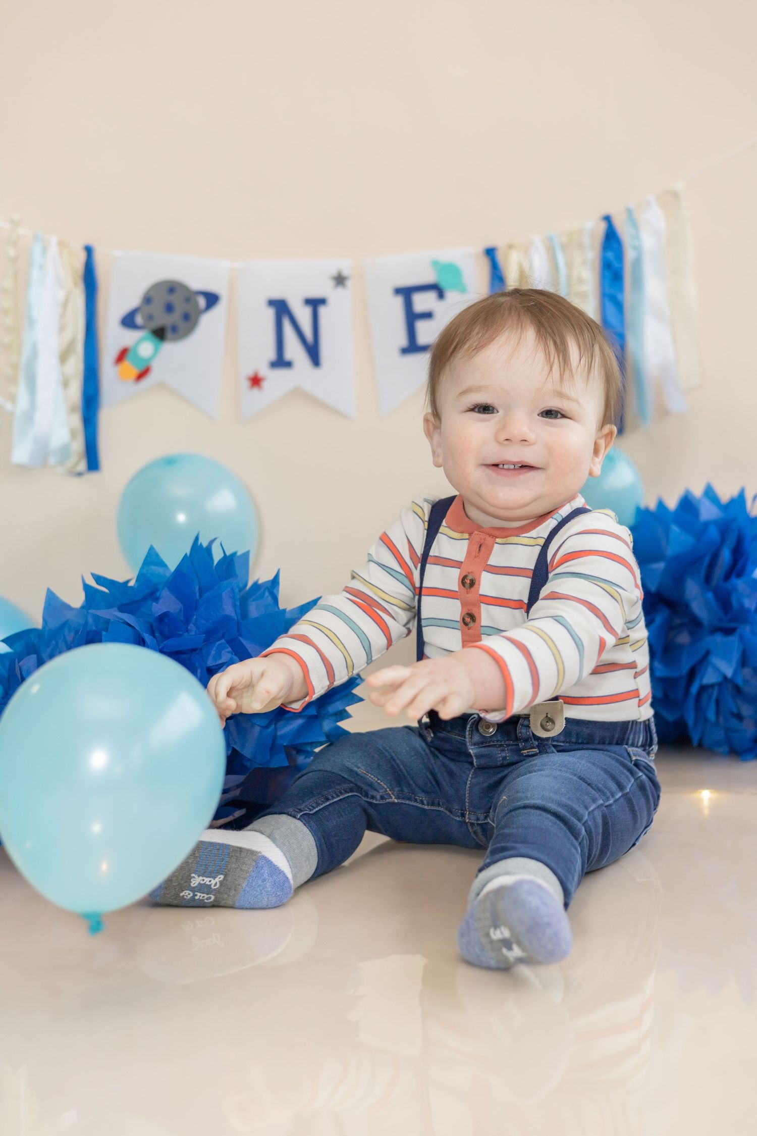 First birthday celebration setup with blue balloons, striped shirt outfit, and space-themed banner decorations in the background.