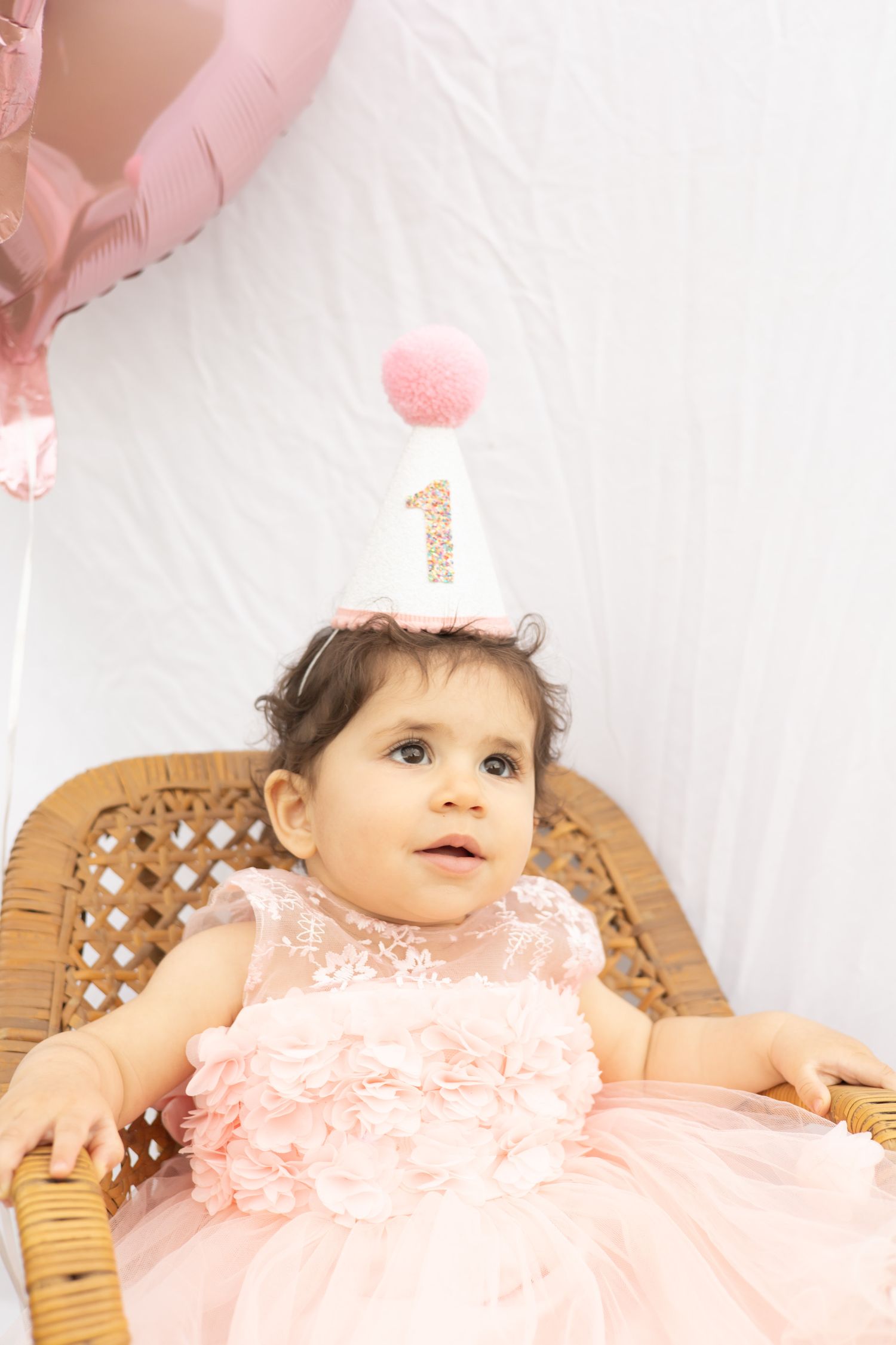 Baby wearing pink lace dress lies on wicker furniture while celebrating first birthday milestone.