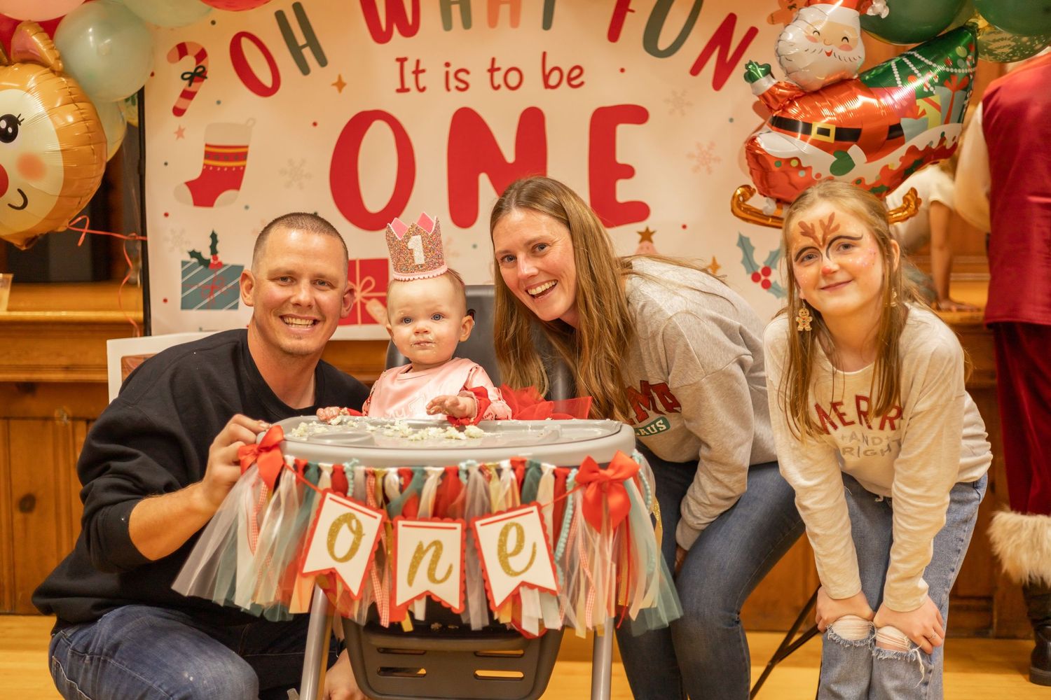 A family celebrates a first birthday party with decorations and a high chair against a festive backdrop.