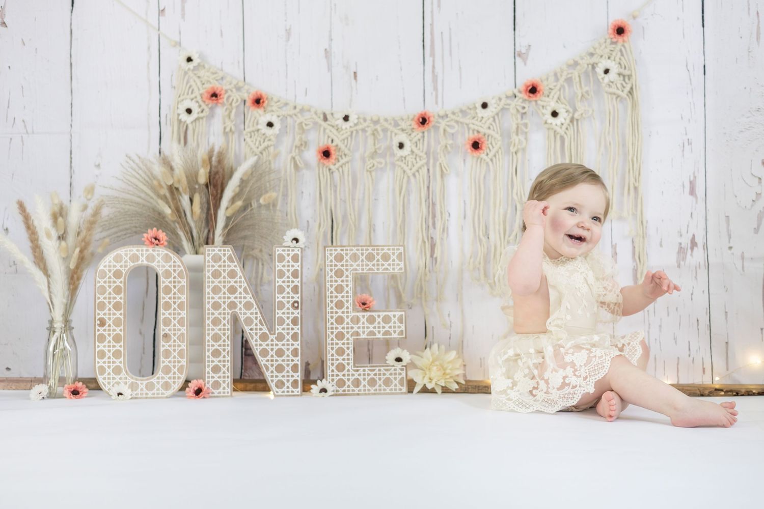Baby in cream lace dress sits beside 'ONE' letters with dried pampas grass and red floral decor on white backdrop.