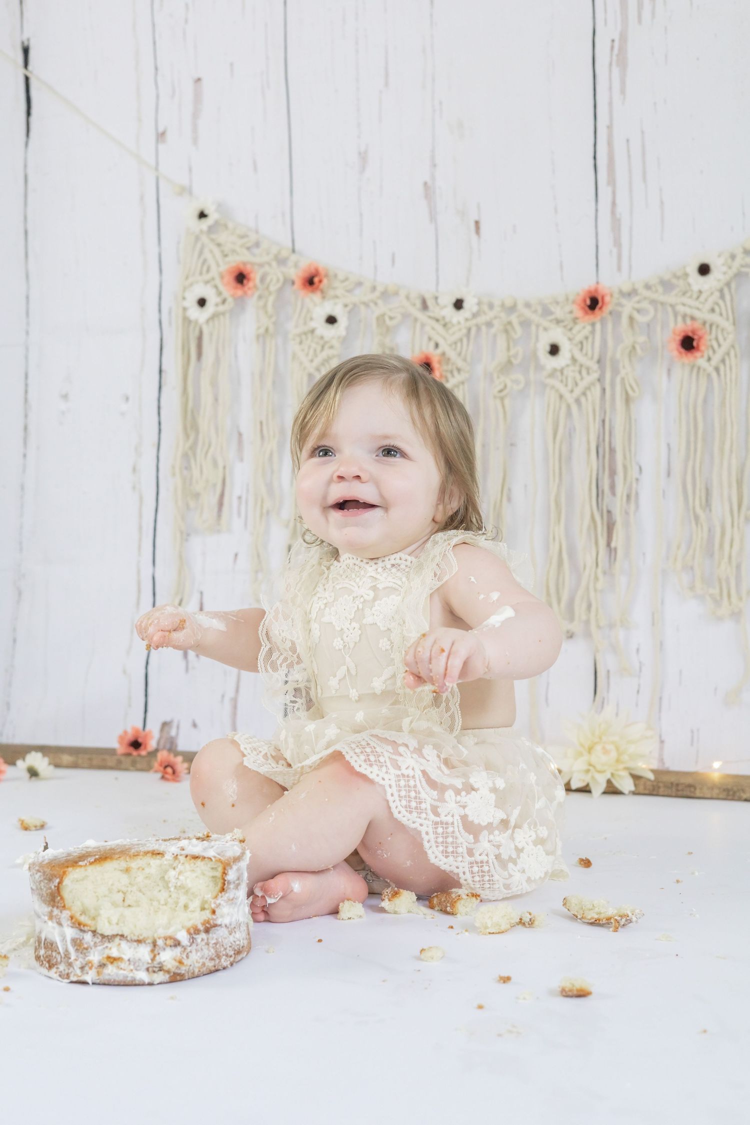 Baby in lace dress with smashed cake during first birthday cake smash photo session against white backdrop.