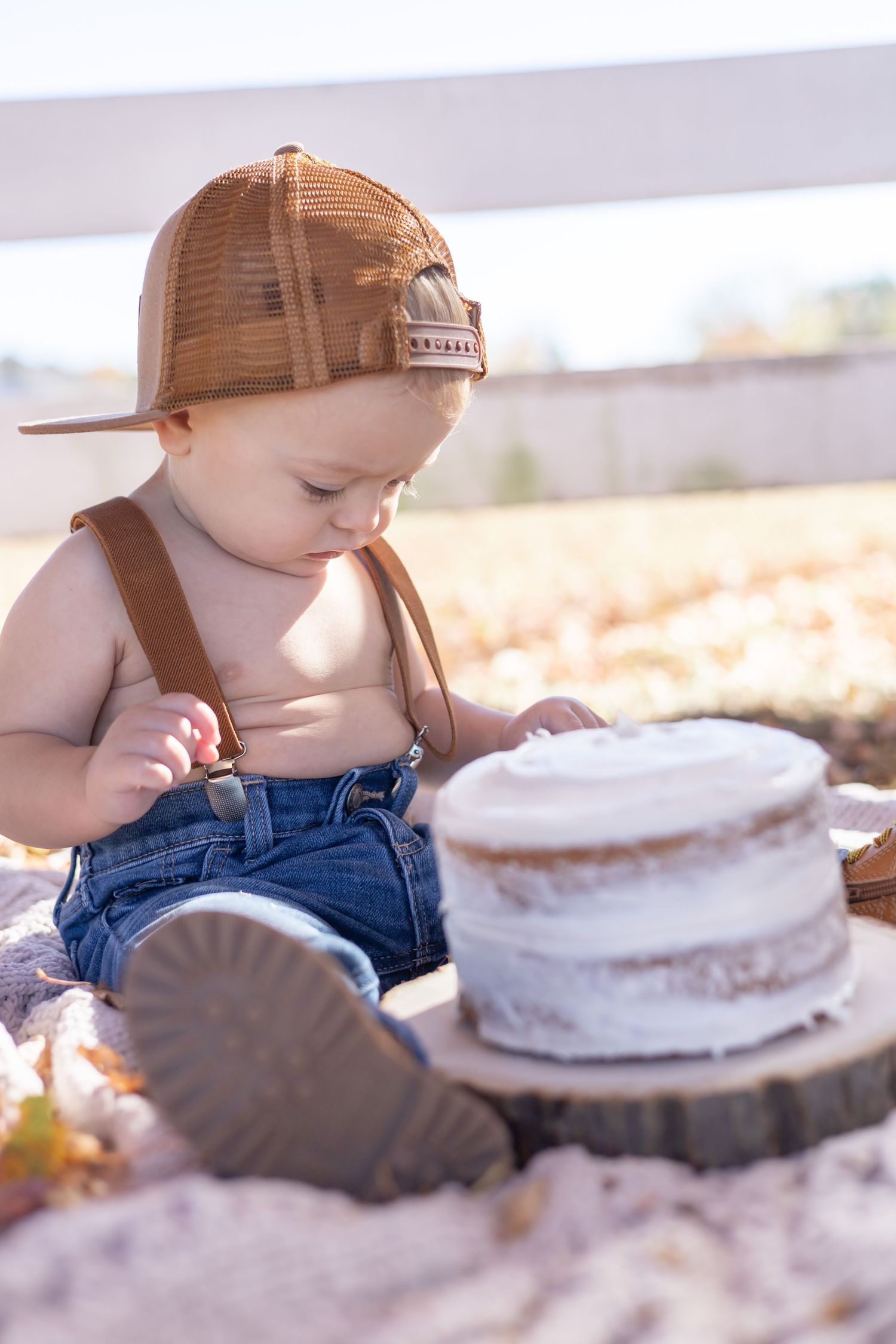 A small child in overalls and a brown cap sits beside a rustic white frosted cake on a wooden slice.