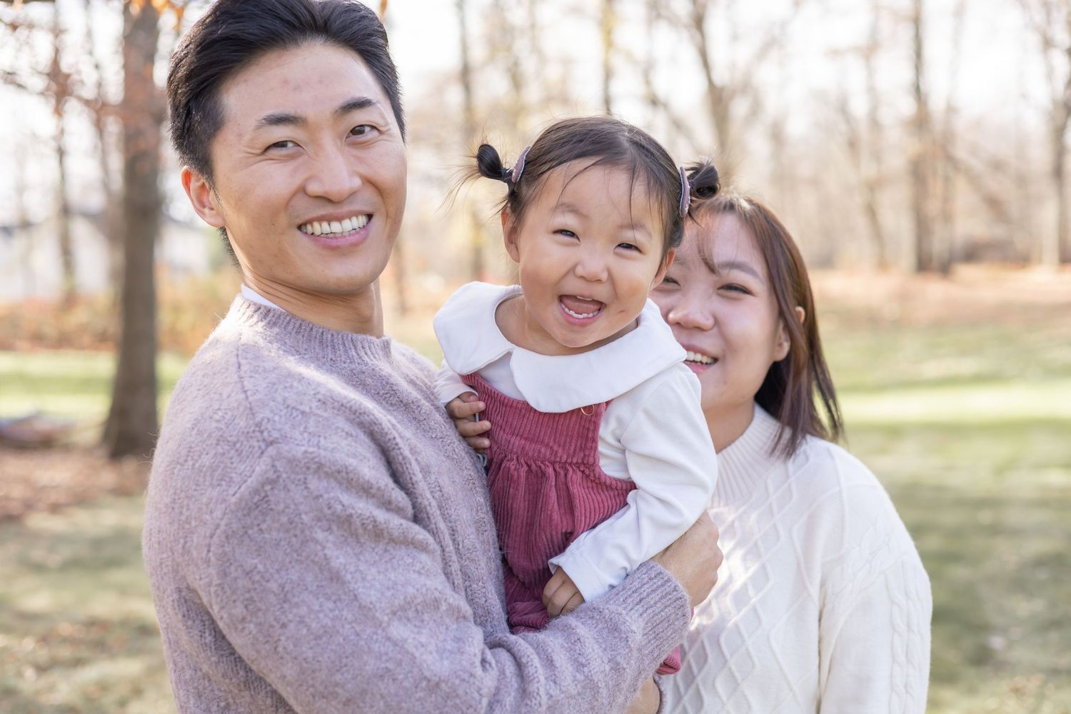 A family stands together outdoors smiling joyfully in an autumn park setting wearing casual attire.