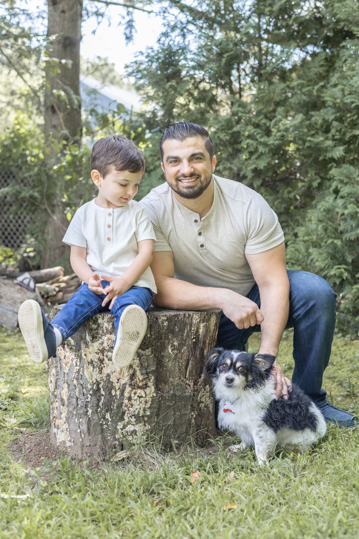 A small black and white dog sits with two people on a tree stump in a sunny backyard garden setting.