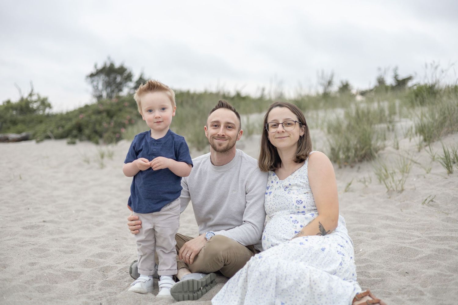 Family sits together on sandy beach dunes during golden hour while wearing coordinated blue and white outfits.