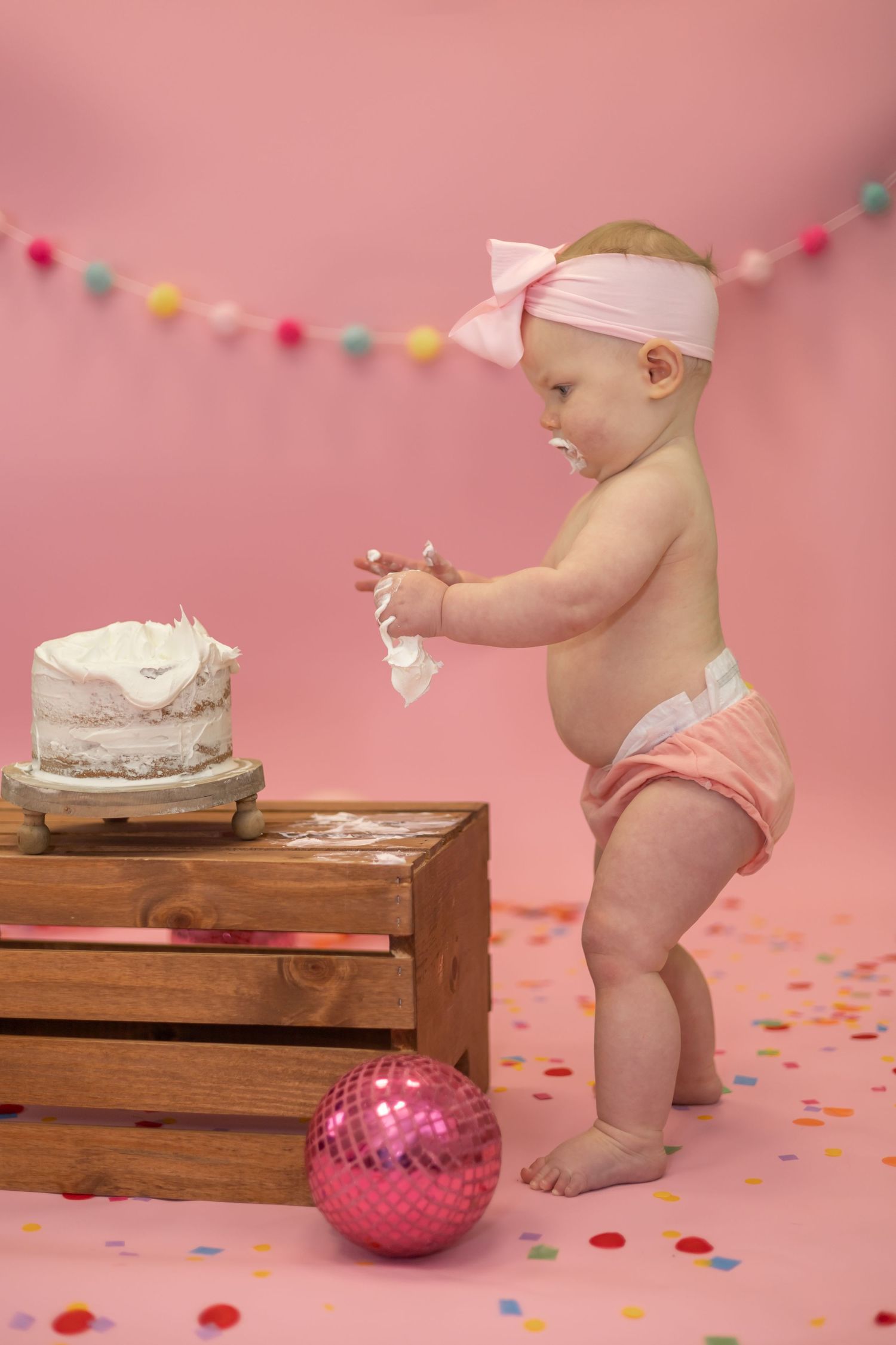 Baby in diaper and headband reaches for cake during first birthday photoshoot with pink background and colorful decorations.