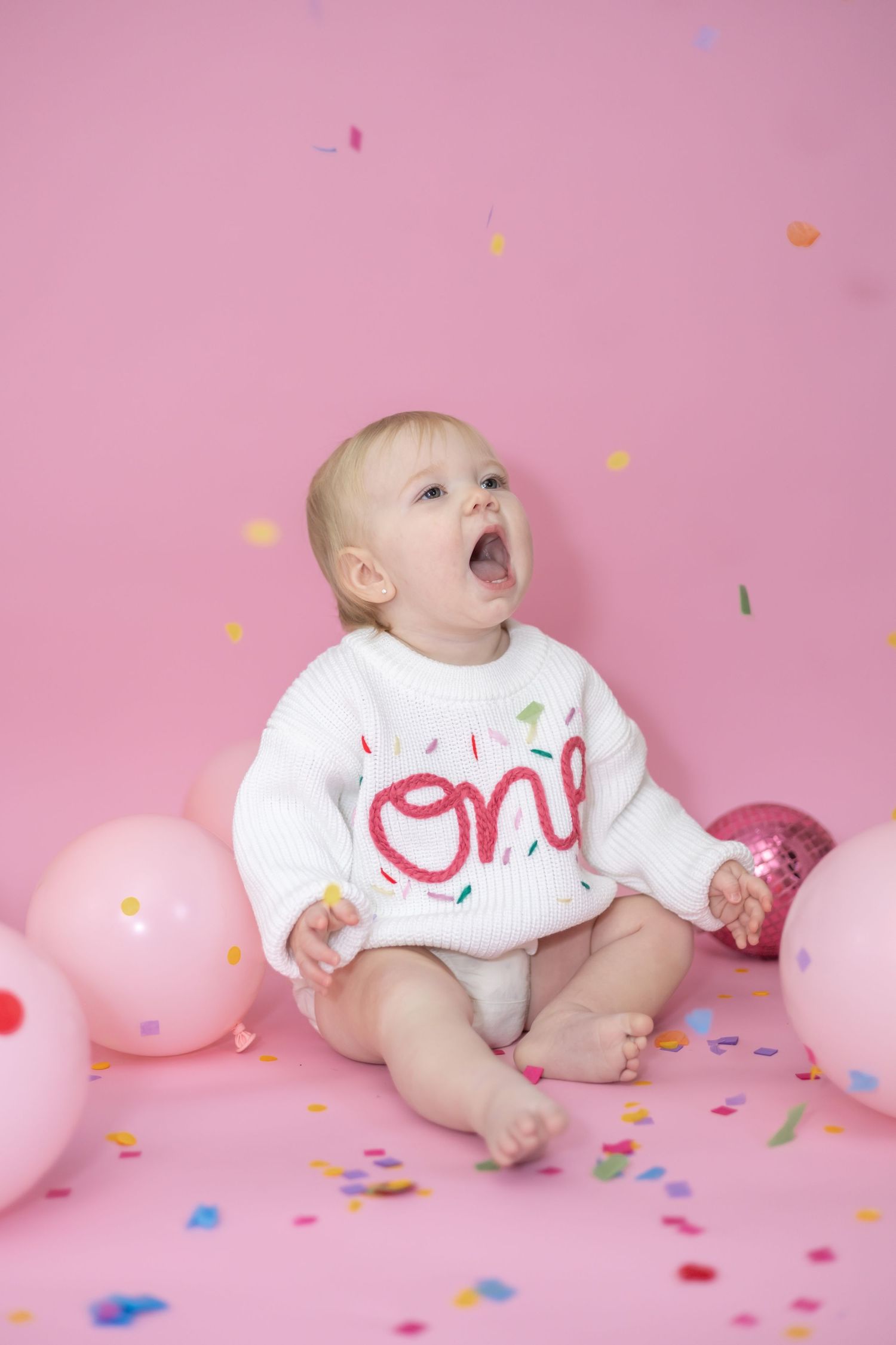 Baby in white onesie lies on pink background with colorful confetti and balloons in playful first birthday photoshoot.