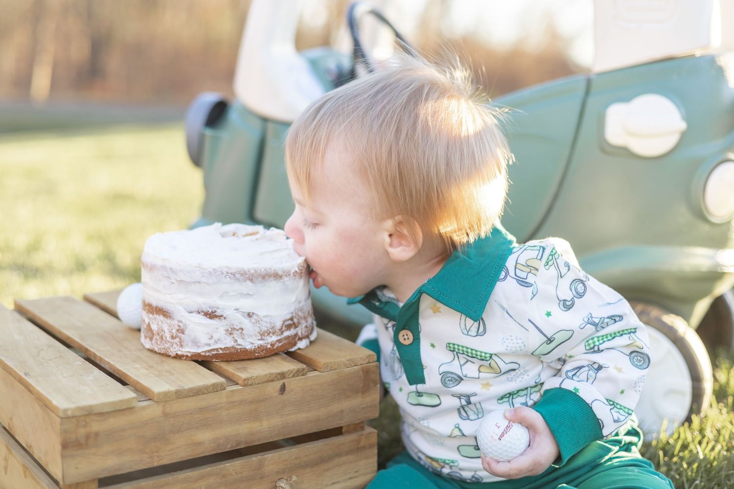 A small child in a patterned outfit leans over to take a bite of a white frosted cake on a wooden surface outdoors.
