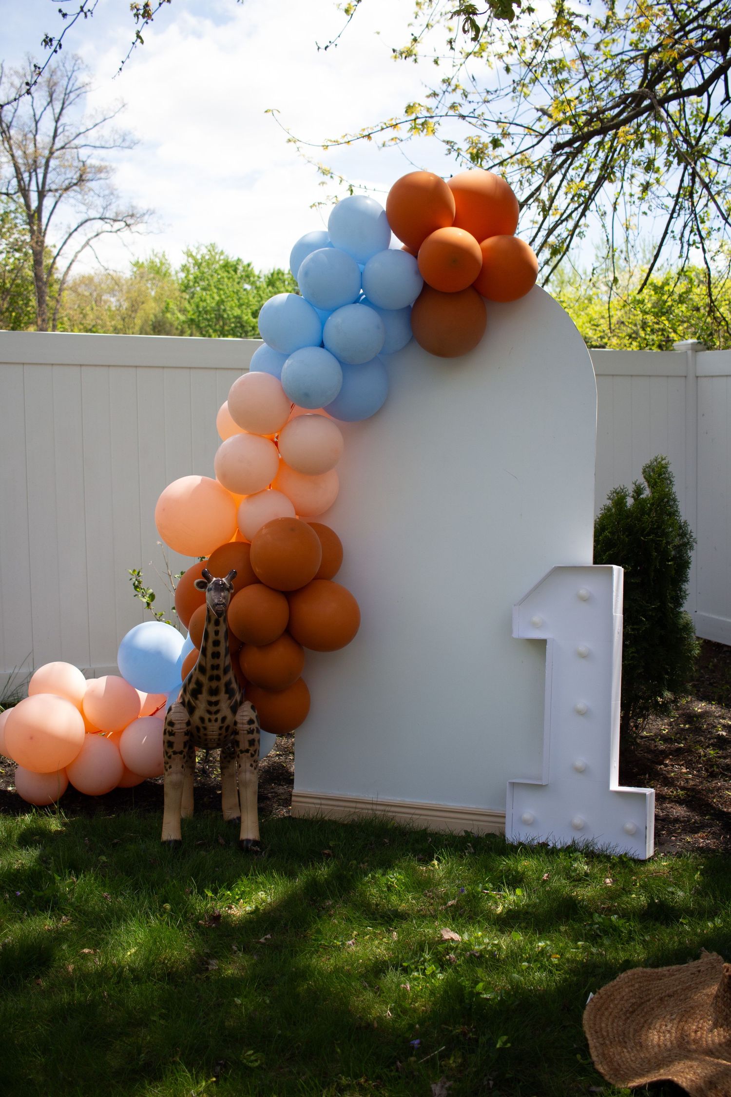 Orange and light blue balloon arch decorates white wall in outdoor garden setting.