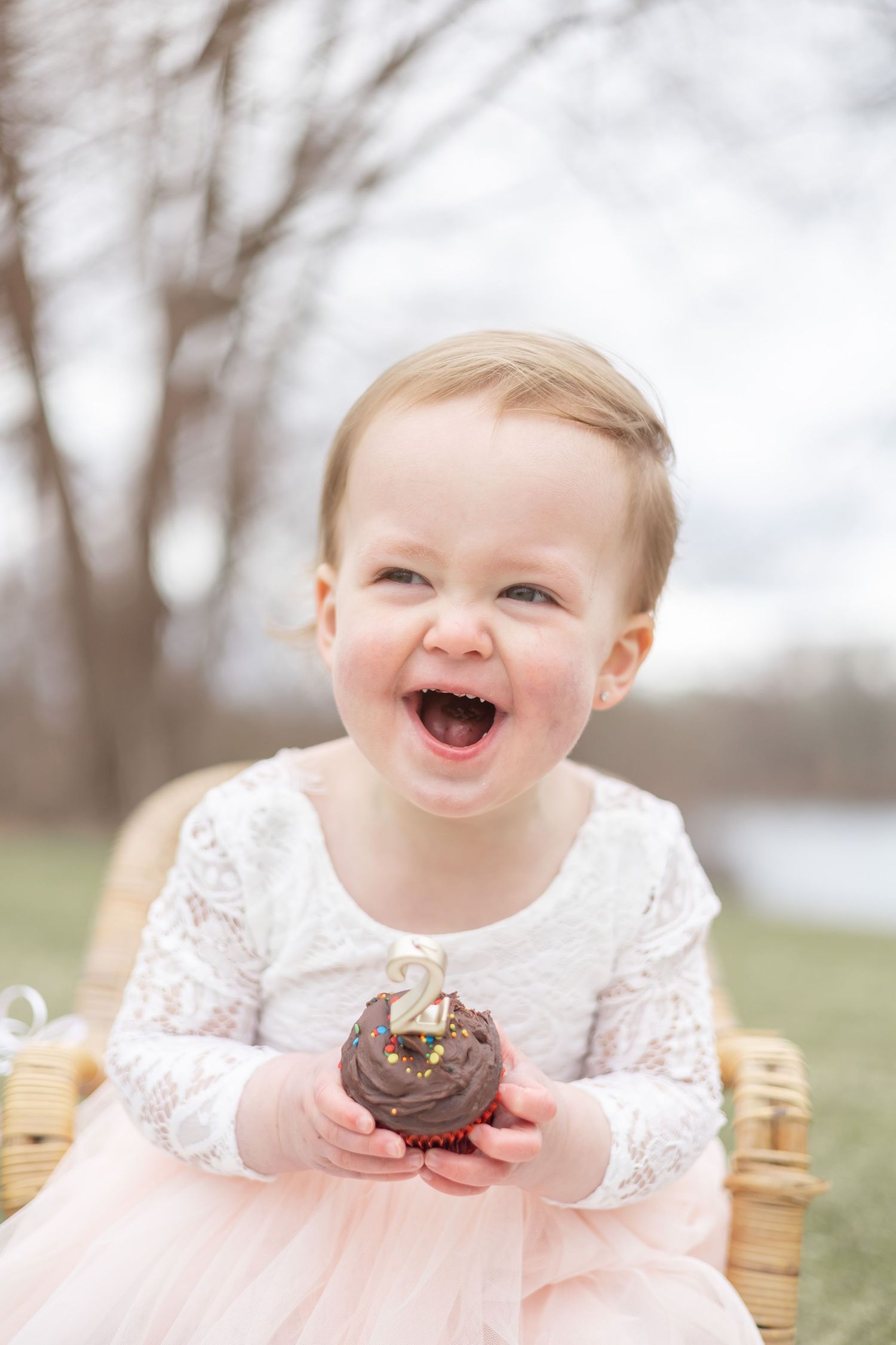 Laughing baby wearing pink tulle dress holds chocolate cupcake while sitting outdoors on rustic chair.