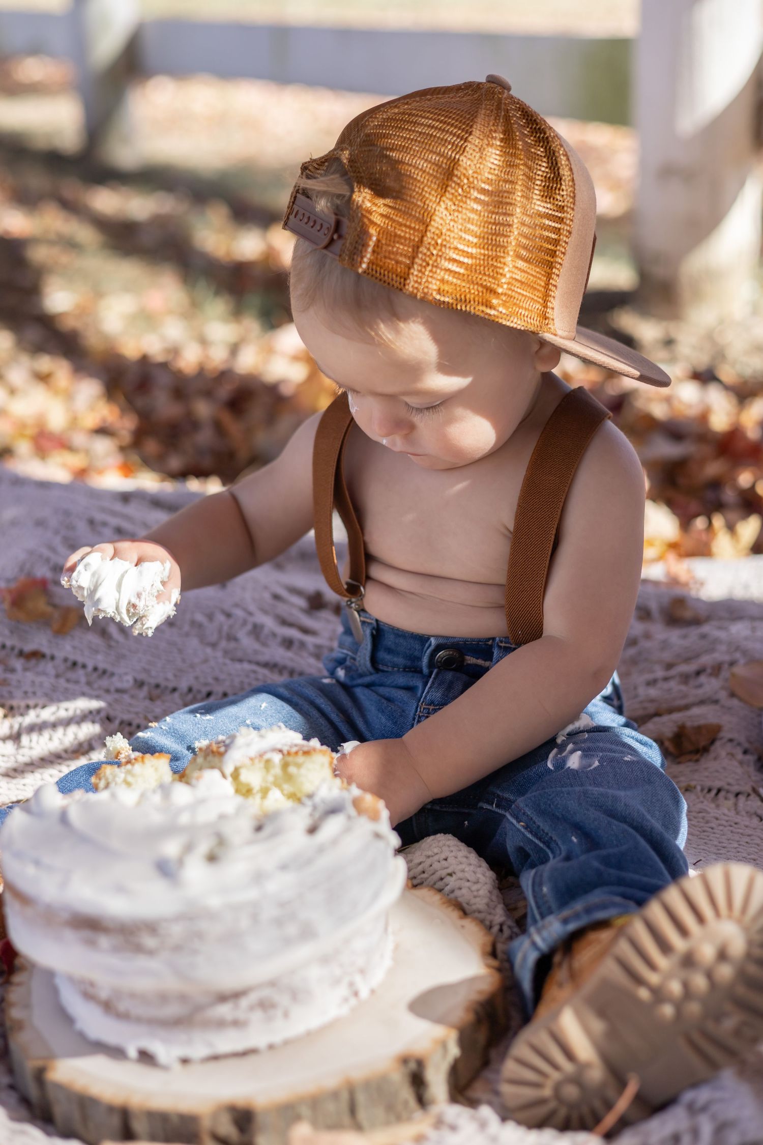 A small child in overalls and a baseball cap sits on a blanket touching a white frosted cake on a rustic wood slice.