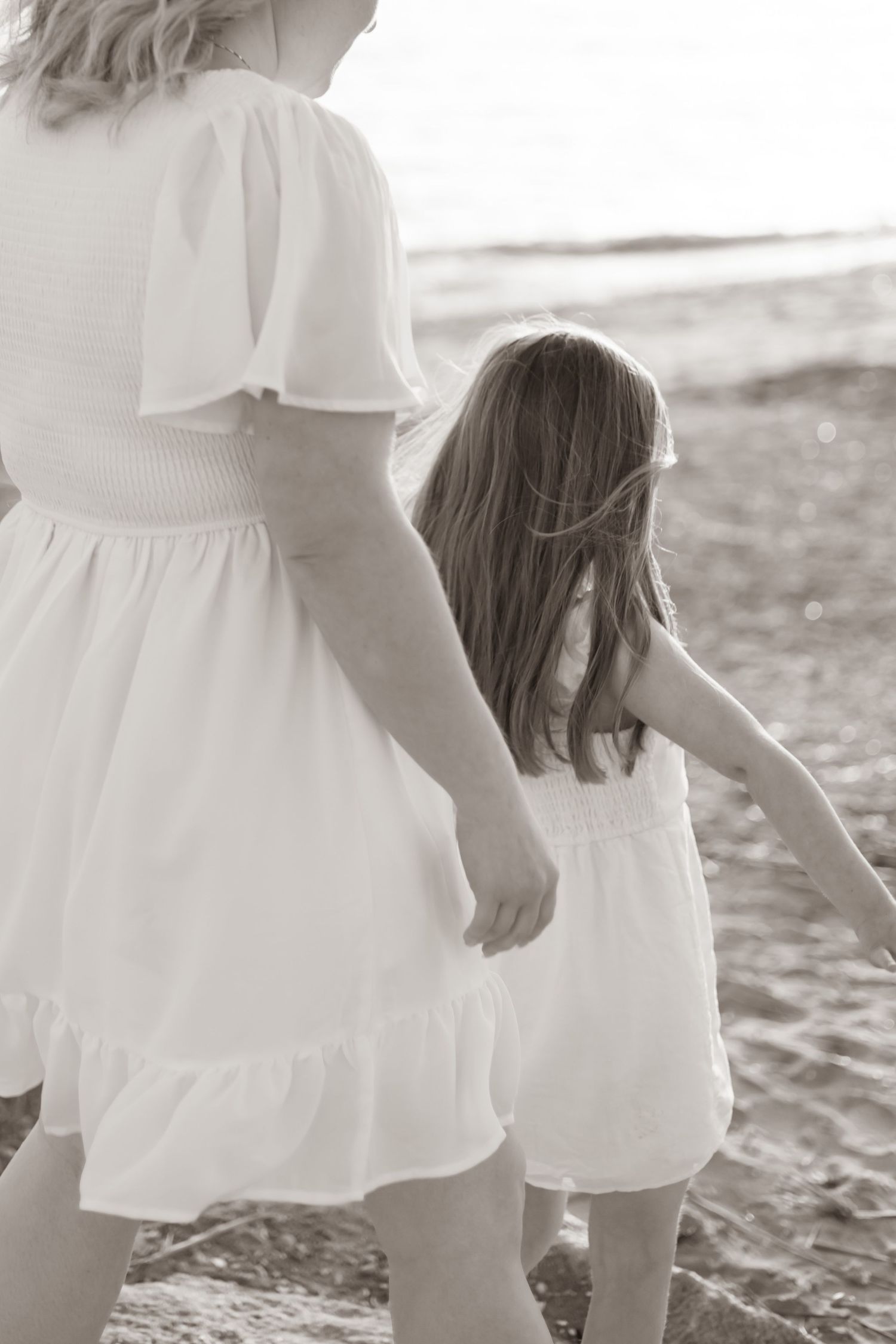 A mother and daughter in white dresses walk together along the beach in a dreamy black and white photograph.